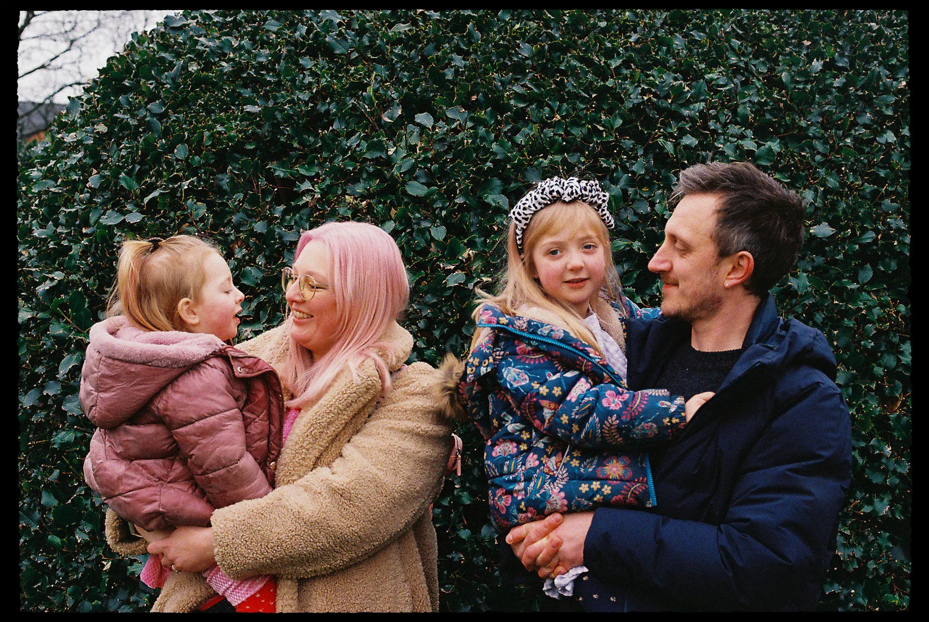 Family outdoors, Mum and Dad and two young girls, smiling and interacting in front of a leafy hedge.