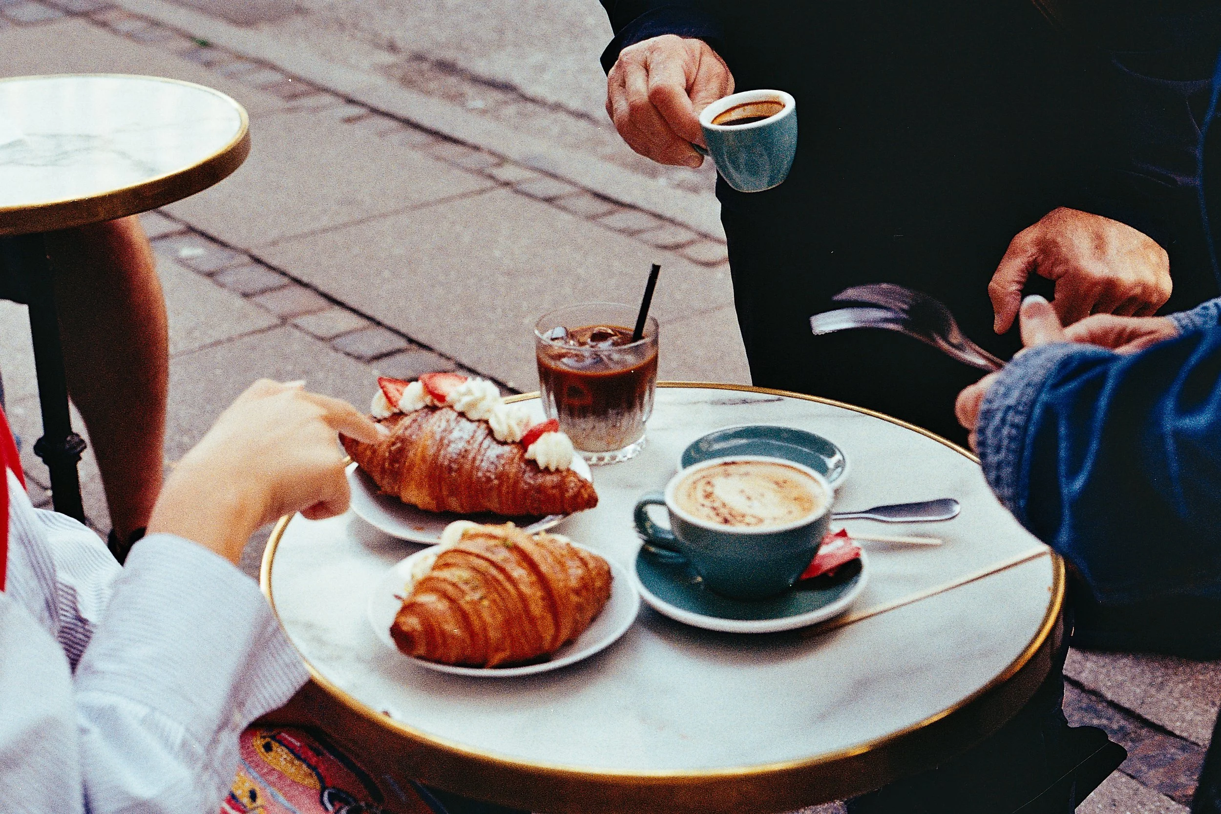 A round marble table at an outdoor cafe with coffee and pastries on it, two people are holding cups, one with a chopstick-stirred drink and another with a small espresso, some pastries include croissants and a strawberry-topped Danish.