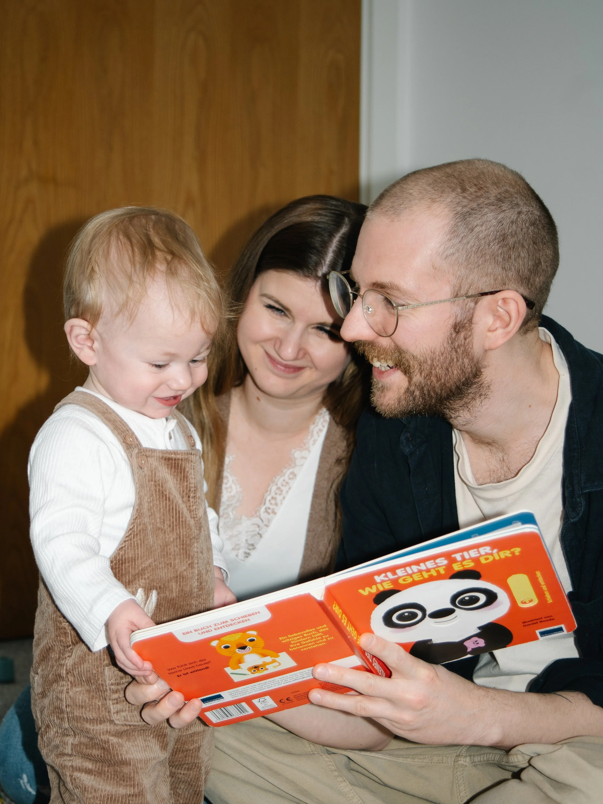 A family with a young child, a woman, and a man sitting together, reading a children's book with a panda bear on the cover. They are smiling and looking at the book.