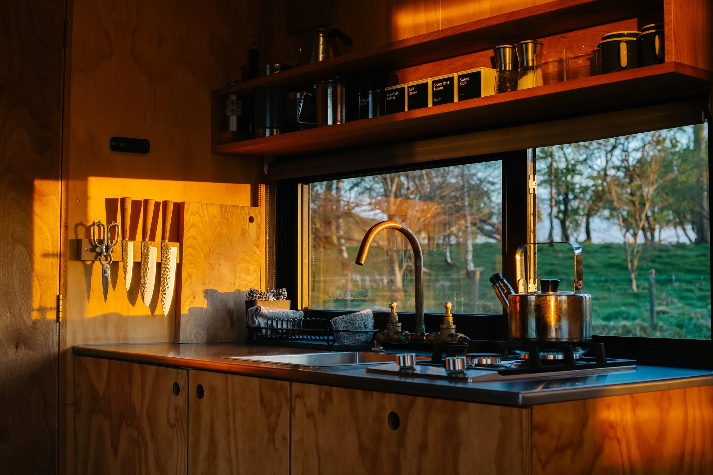 Cozy kitchen with a window overlooking a grassy landscape with trees, a wooden countertop, hanging knives, and shelves with kitchenware in warm sunlight.