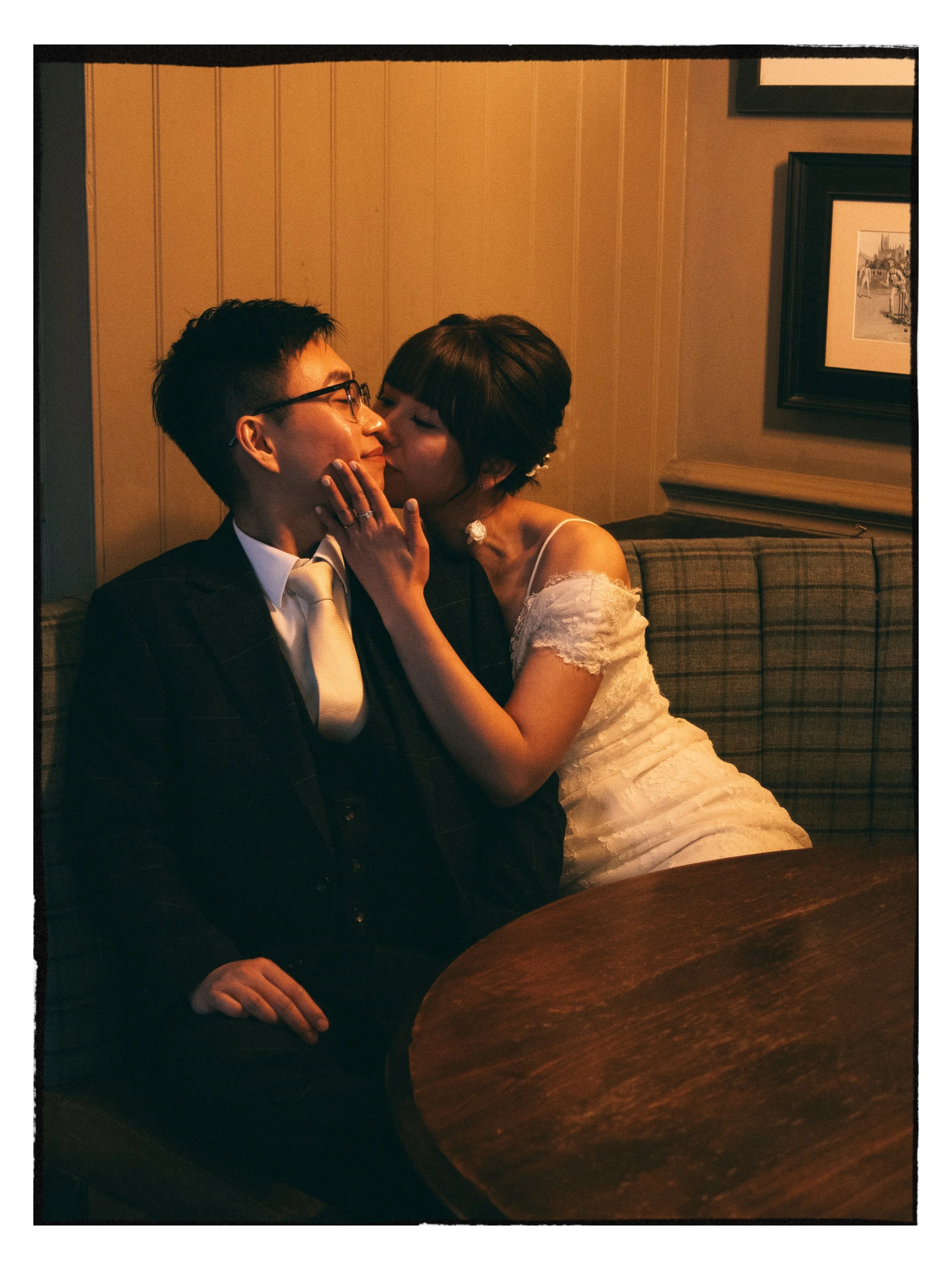 A bride is kissing her groom on the cheek while touching his face. The groom is wearing glasses and a suit, and they are sitting on a checkered couch.
