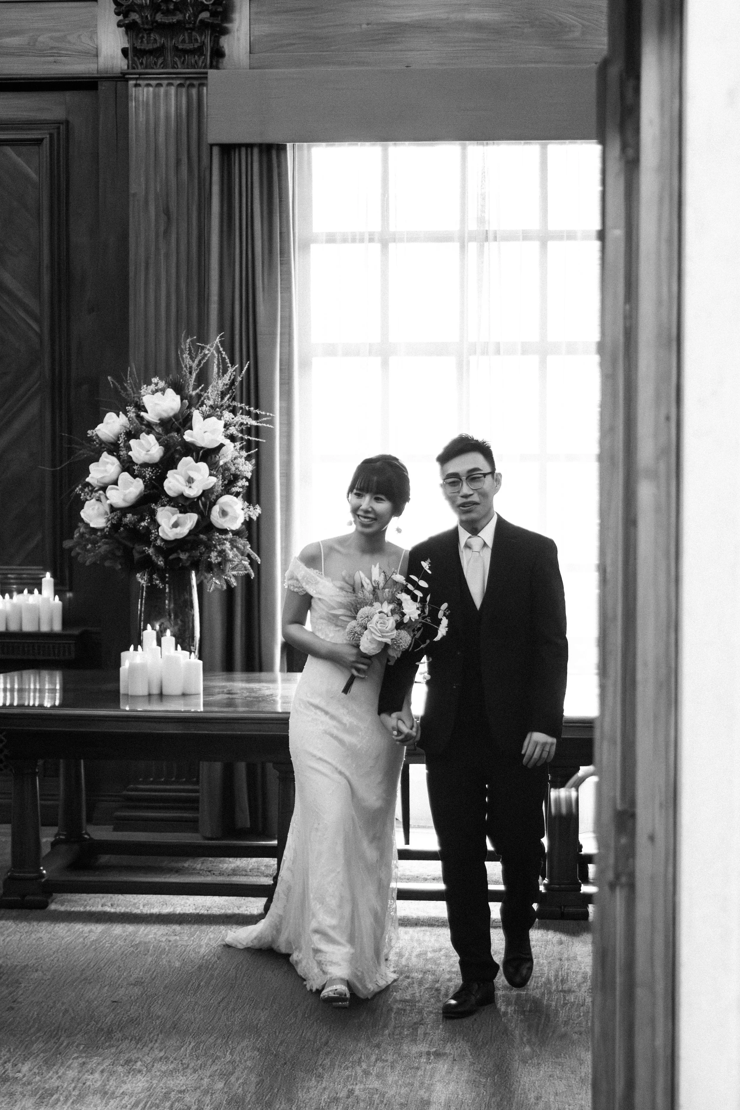 A black-and-white photo of a bride and groom walking hand in hand in a room with large window. The bride is in a wedding dress holding a bouquet, and the groom is in a suit. A large floral arrangement and candles are on a table nearby.