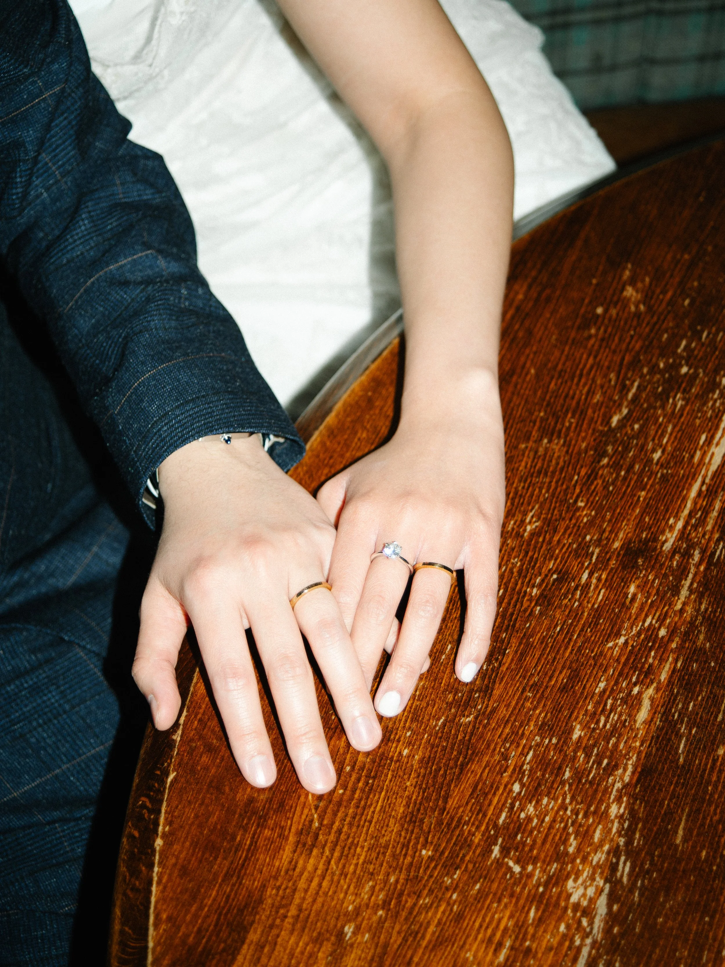 Close-up of a couple holding hands on a wooden table, showing wedding rings and engagement ring.