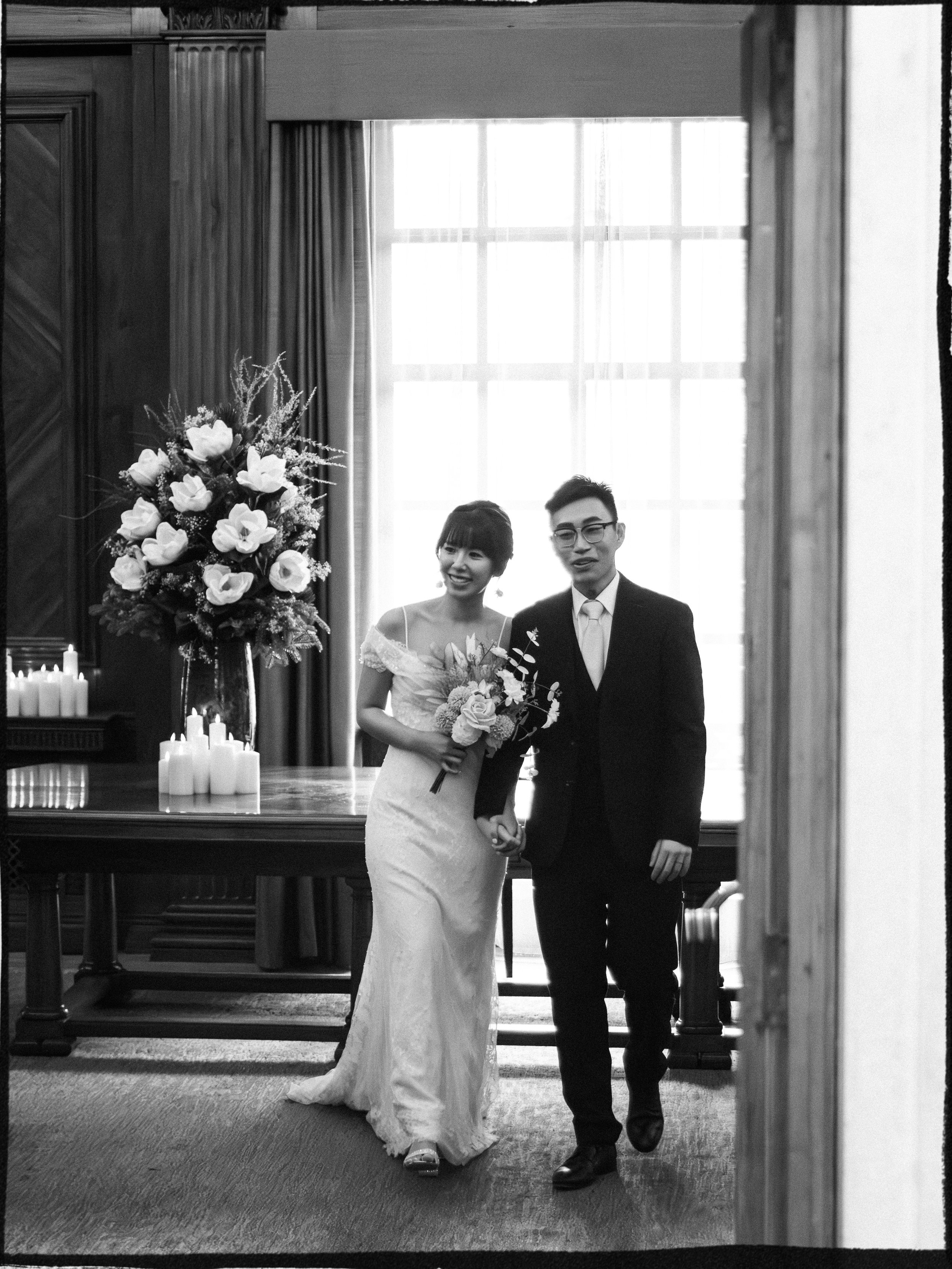 A bride and groom holding hands and smiling in a formal indoor setting with a large window, floral arrangements, and candles.