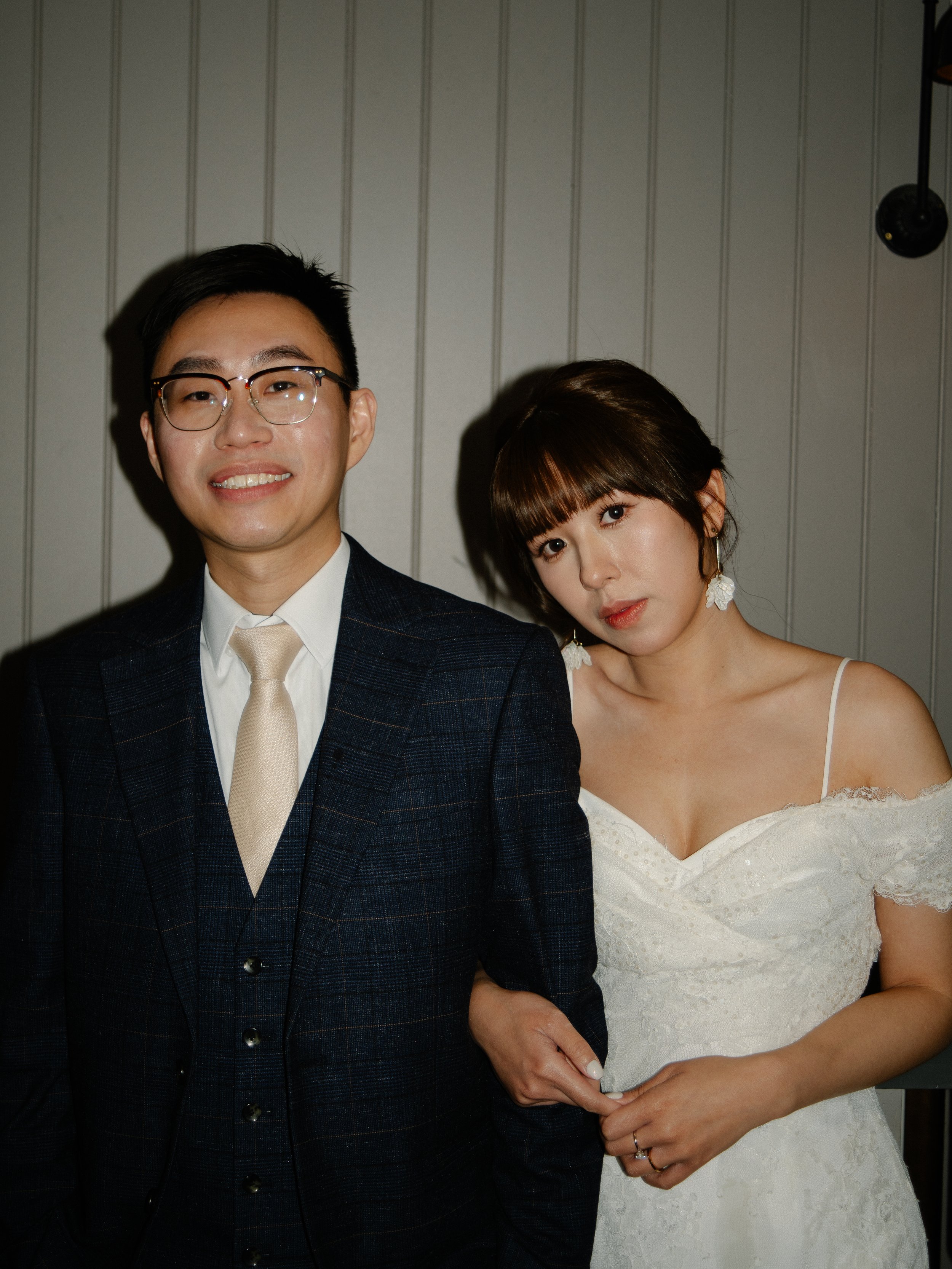 A man in glasses and a woman in a white dress standing together indoors against a wood-paneled wall.