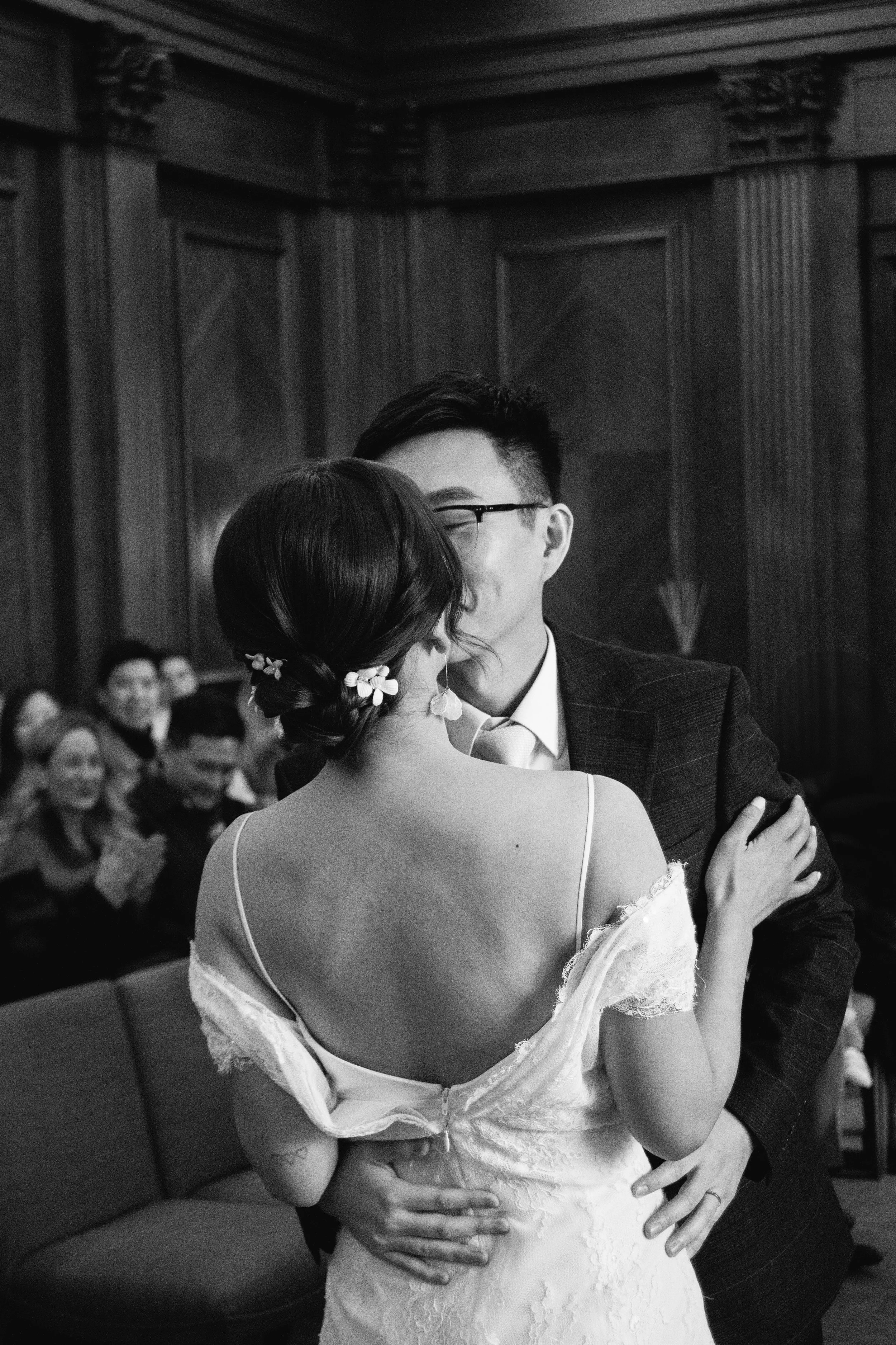 A black and white photo of a newly married couple sharing their first dance at a wedding, surrounded by seated guests in a wood-paneled room.