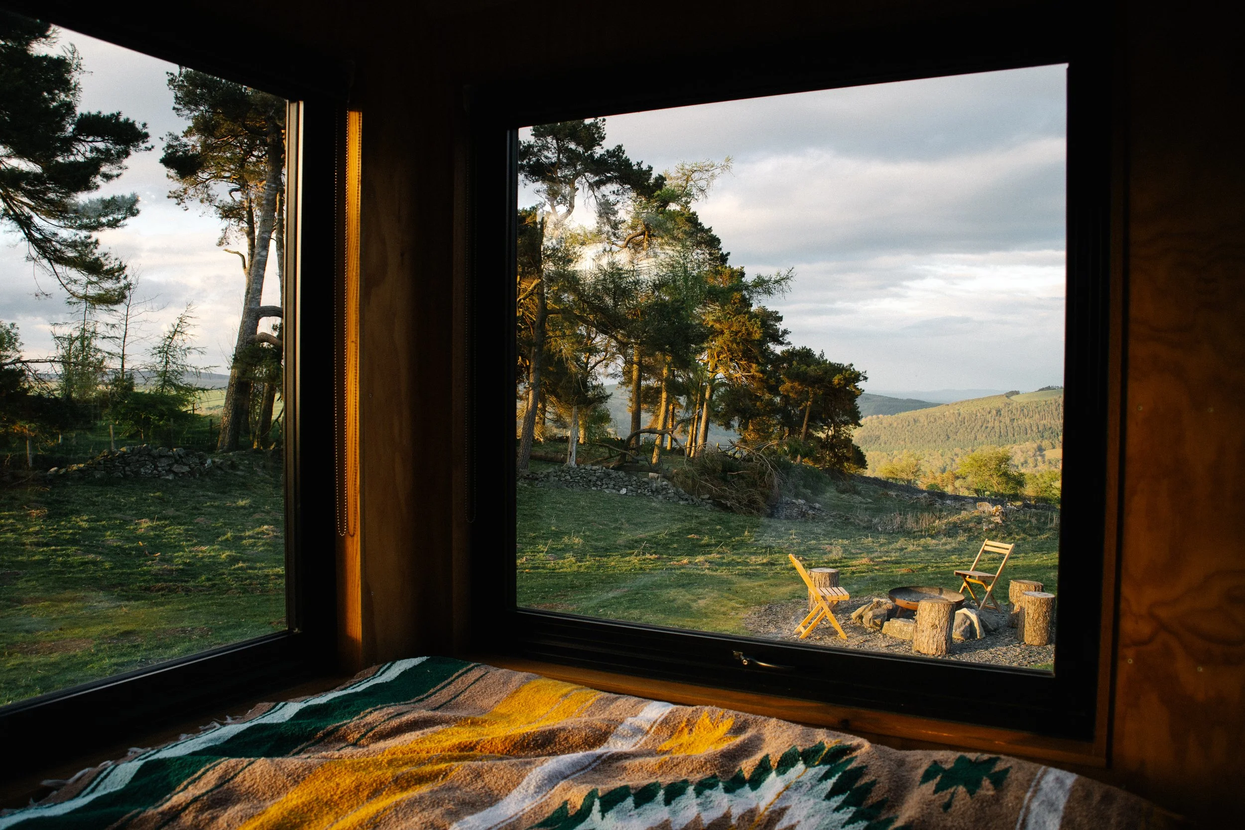 View of a forested landscape through large corner window, with outdoor chairs and a fire pit on a grassy area in the evening sunlight.
