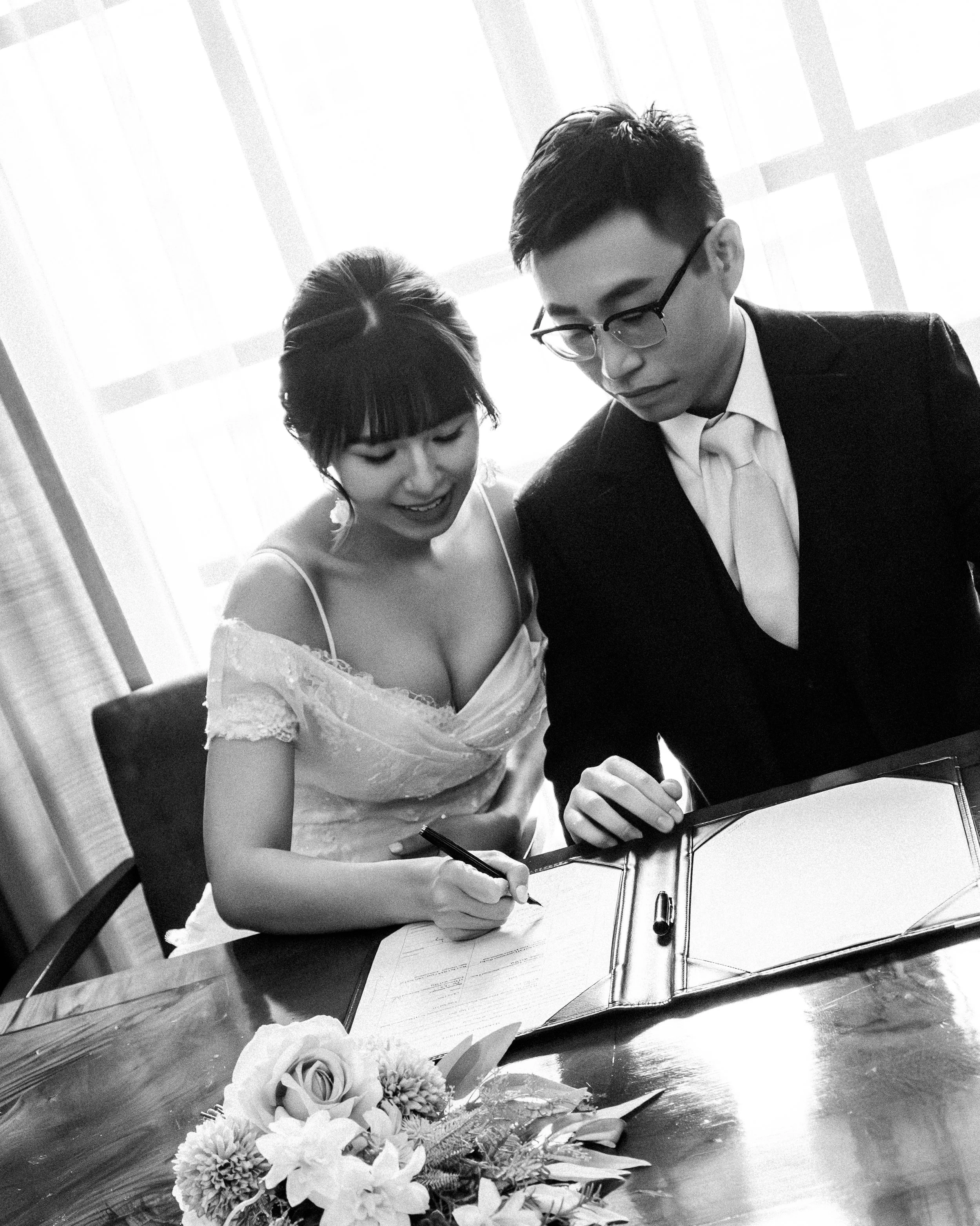 A bride and groom signing a marriage certificate at a table, with a flower arrangement in front of them.