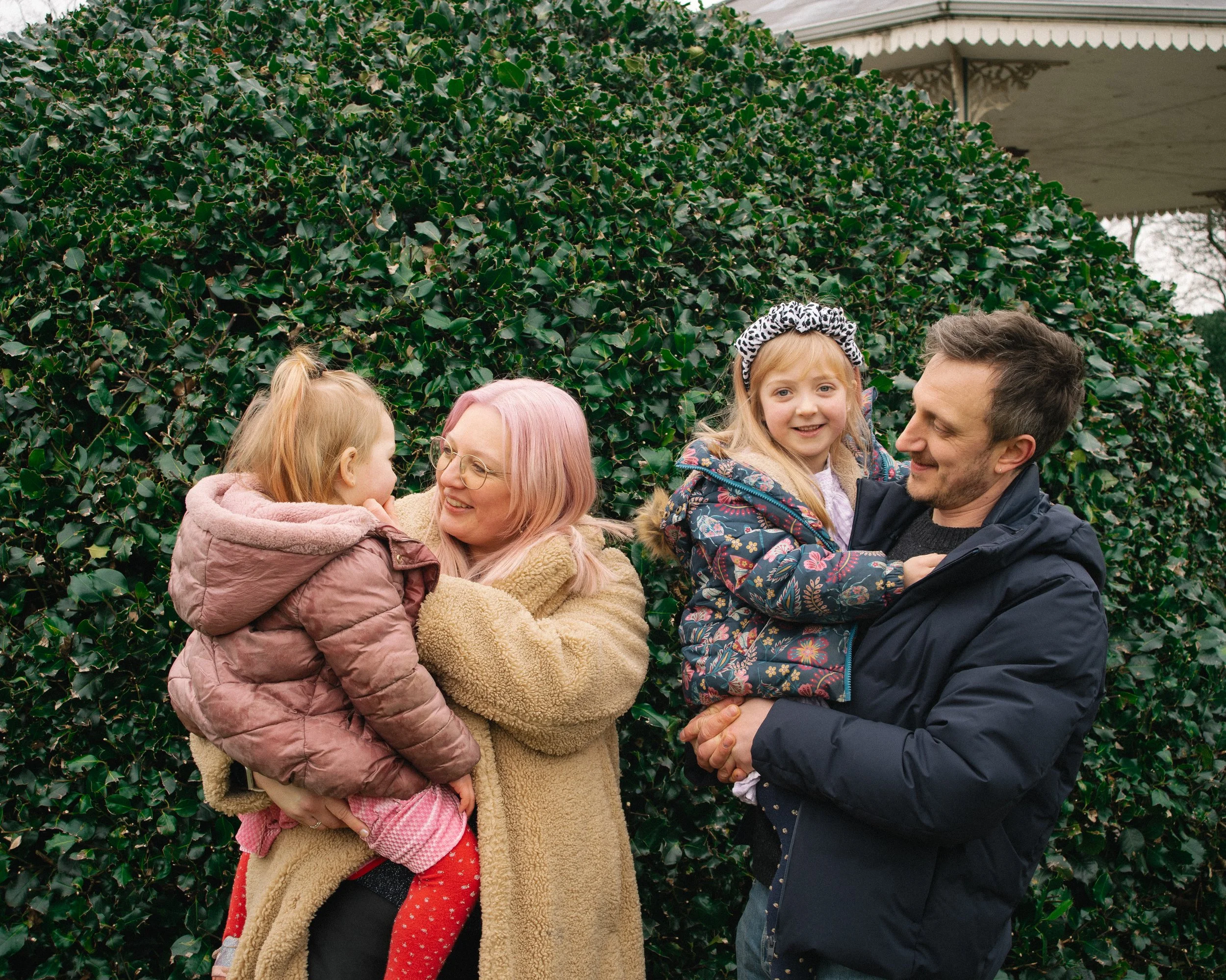 A family of four, two adults and two children, smiling and enjoying time outdoors near a dense hedge.