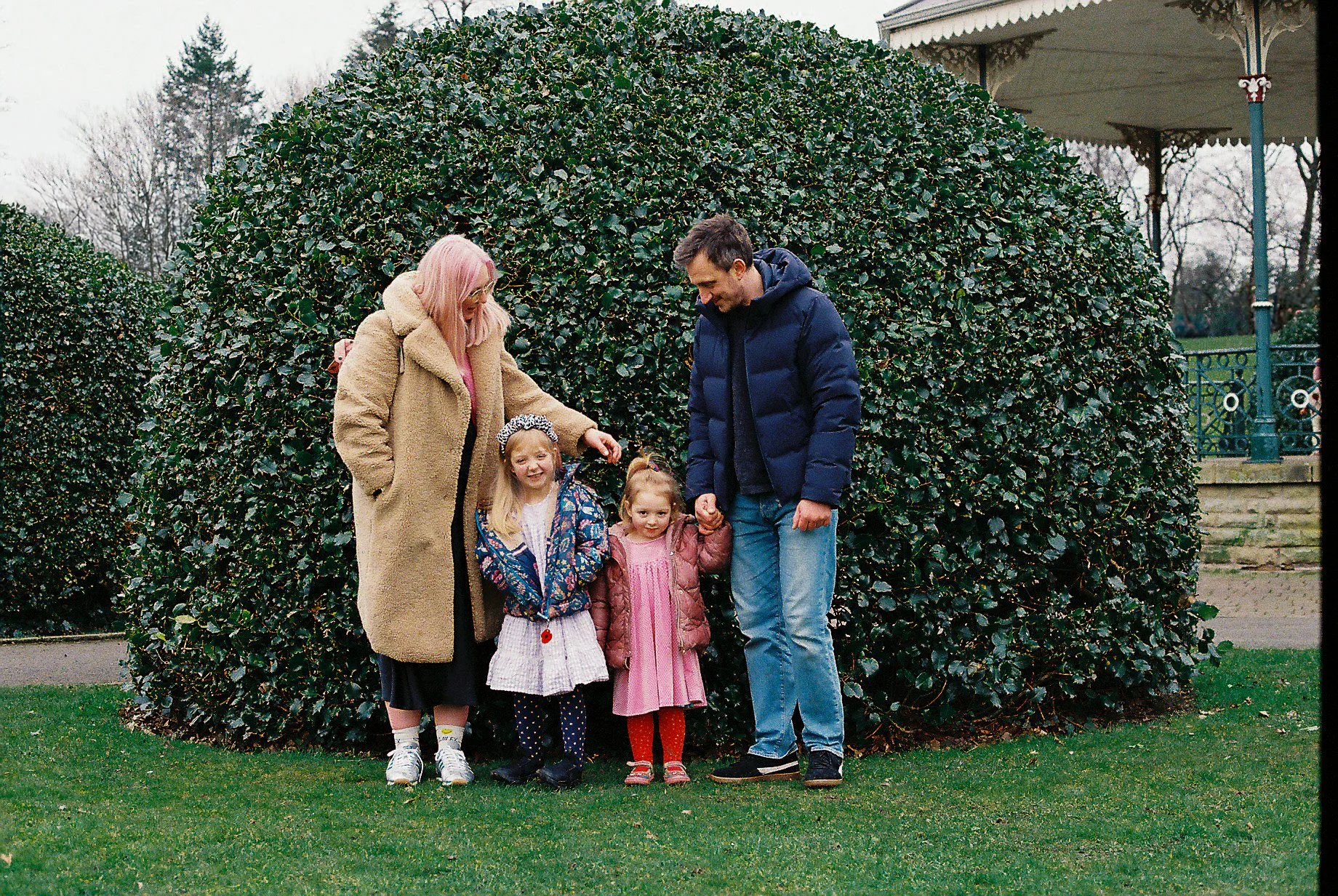 Family outdoors in front of large neatly trimmed bushes, with two adults and three children smiling and holding hands on a cloudy day.