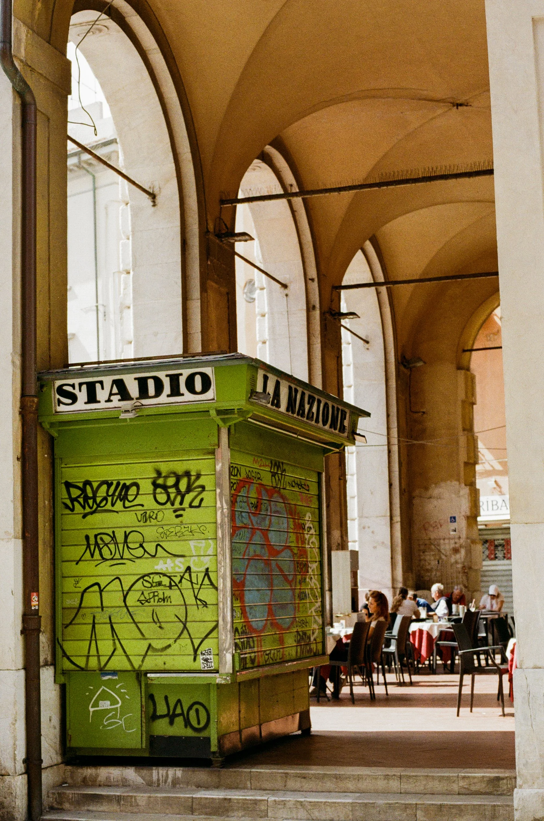 Empty green and white kiosk with graffiti, labeled 'Stadio La Nazione,' set against an arched colonnade with outdoor dining tables and chairs, and people dining.