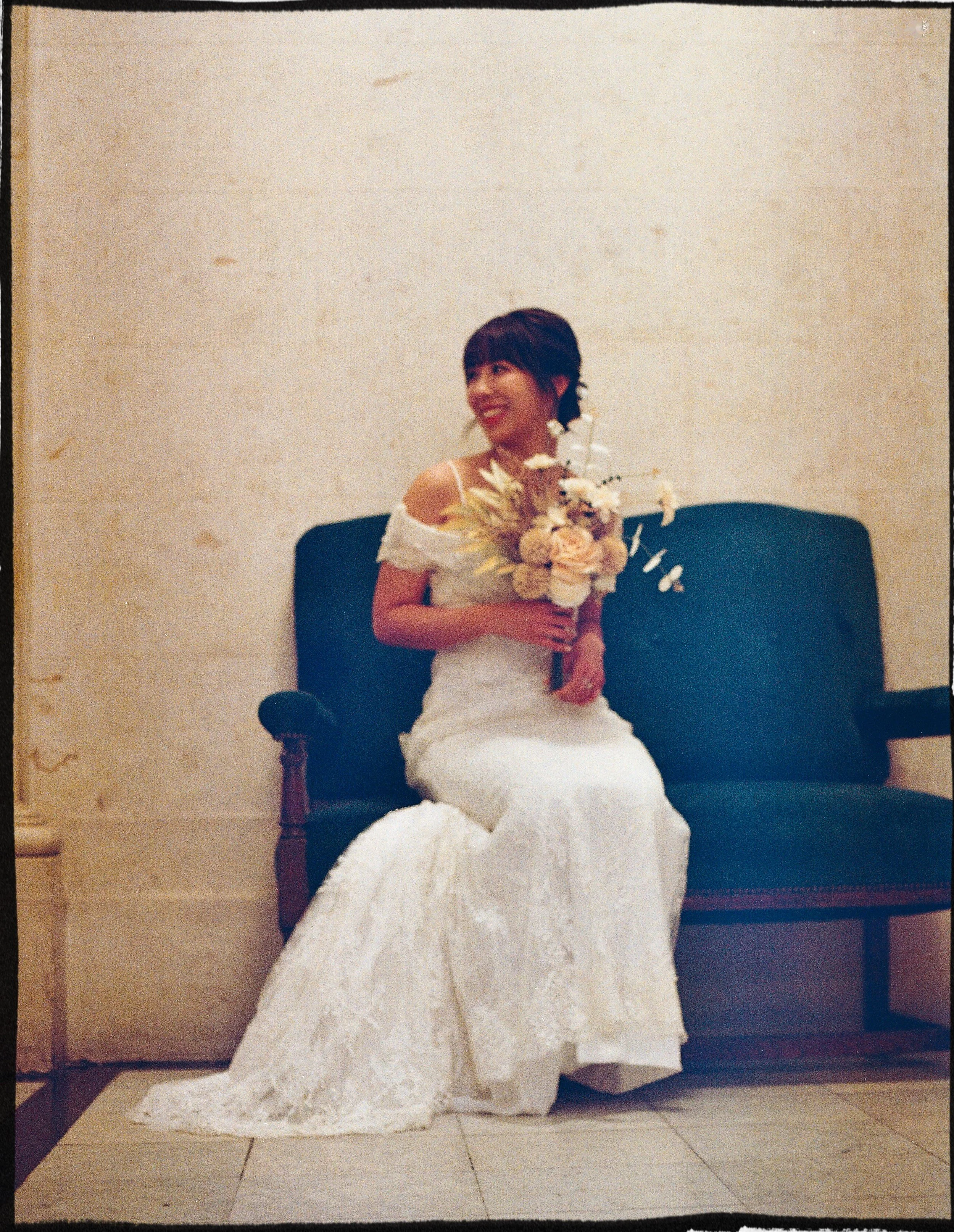 A bride in a white bridal gown sitting on a dark green vintage-style sofa holding a bouquet of flowers, smiling, with a beige stone wall background.