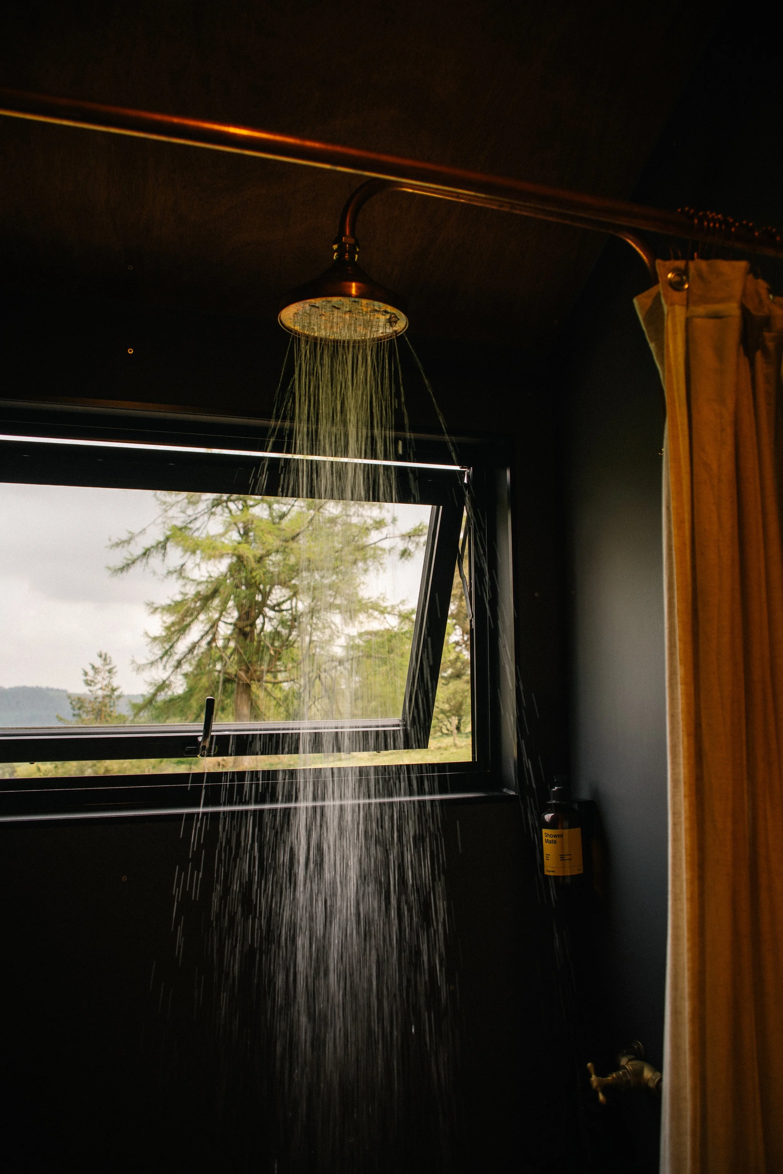 Shower with water running inside a dark room, with a window showing trees and cloudy sky outside, and a yellow curtain to the right.