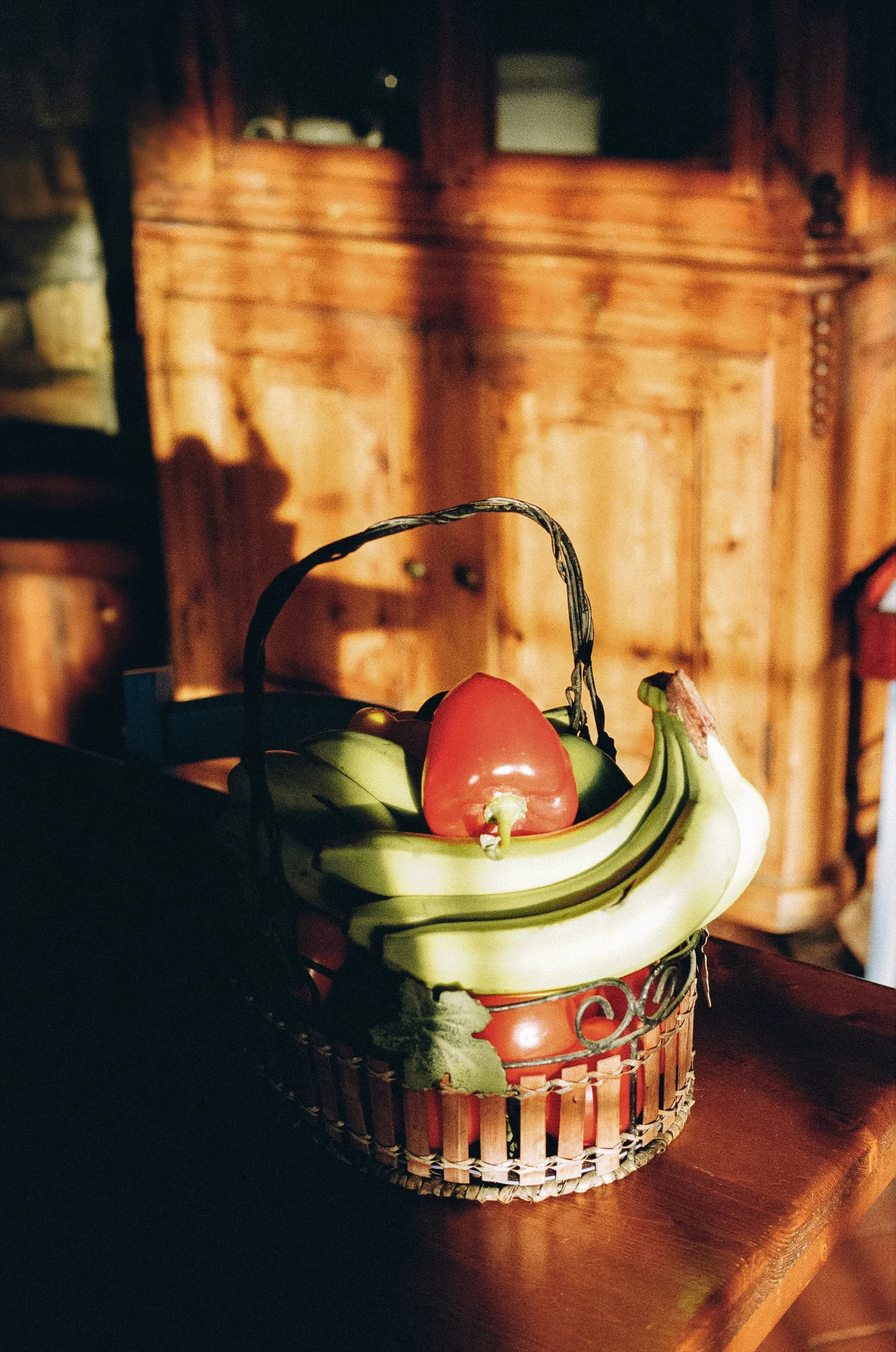 A basket of artificial fruits including bananas, a tomato, and a red bell pepper, placed on a wooden surface with a wooden cabinet in the background.