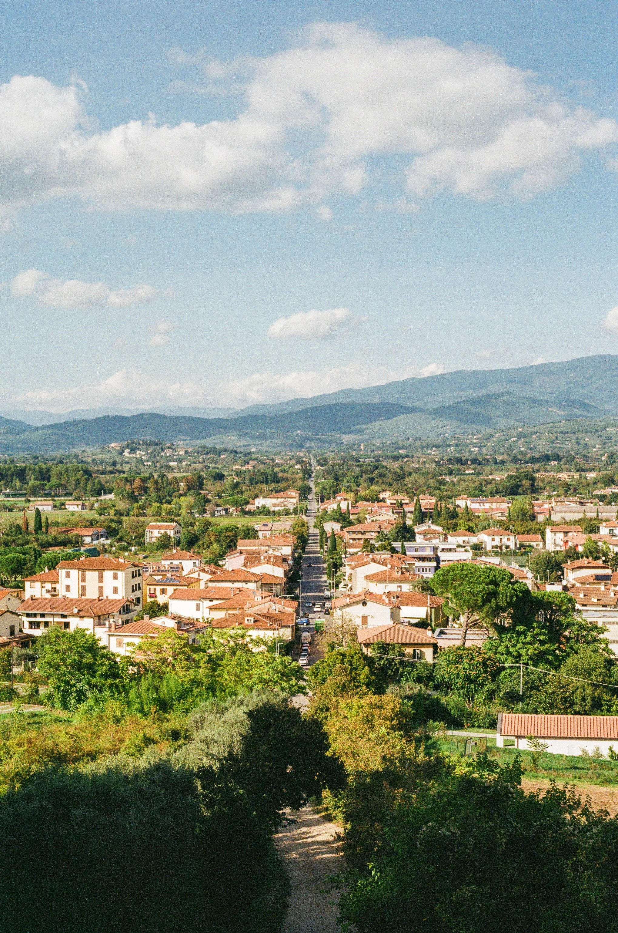 A scenic view of a suburban neighborhood with red-tiled roofs, green trees, and a straight road leading towards distant mountains under a partly cloudy sky.