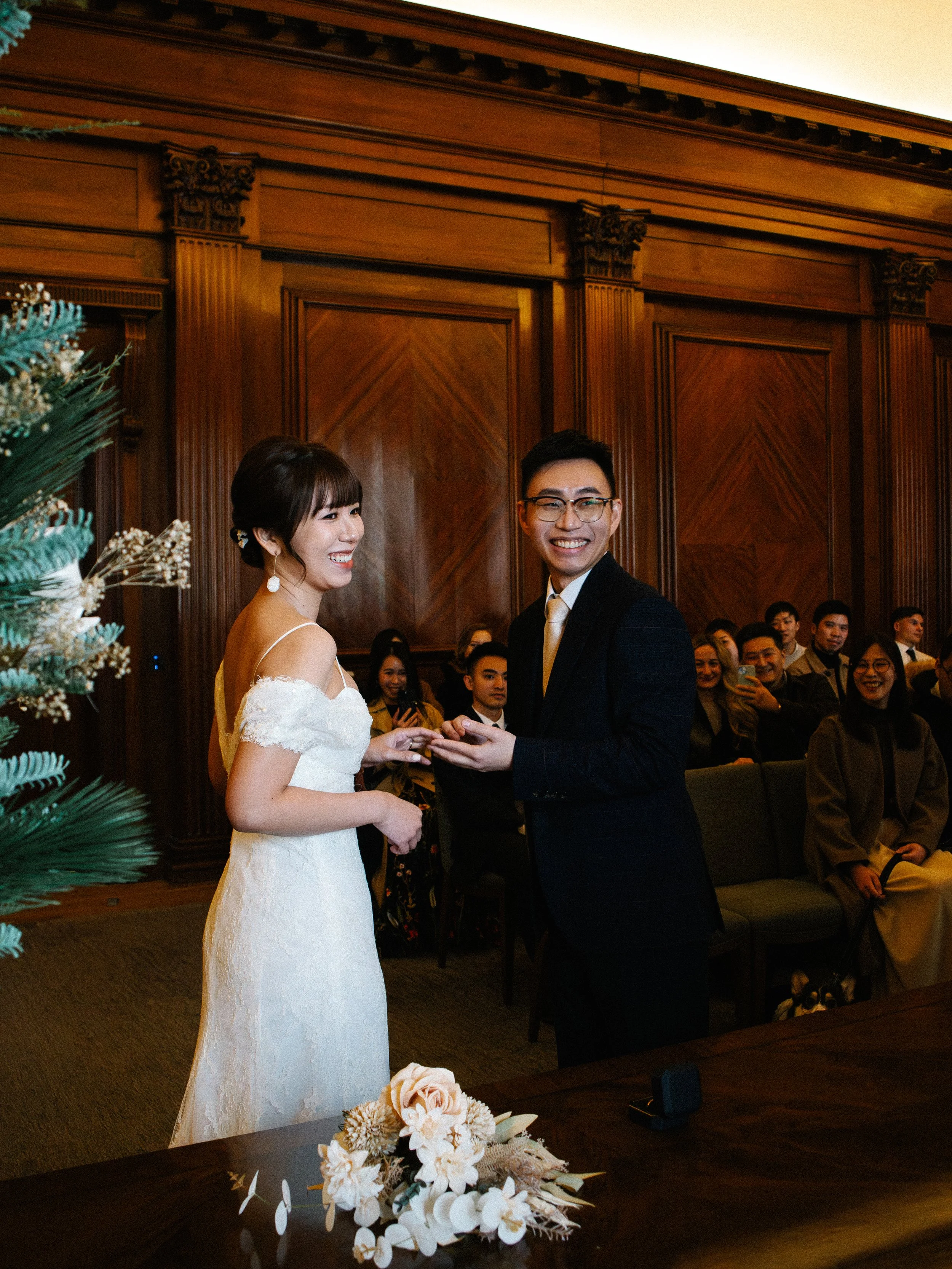 A bride and groom exchange wedding rings at their wedding ceremony in a wooden-paneled room with seated guests watching and smiling.