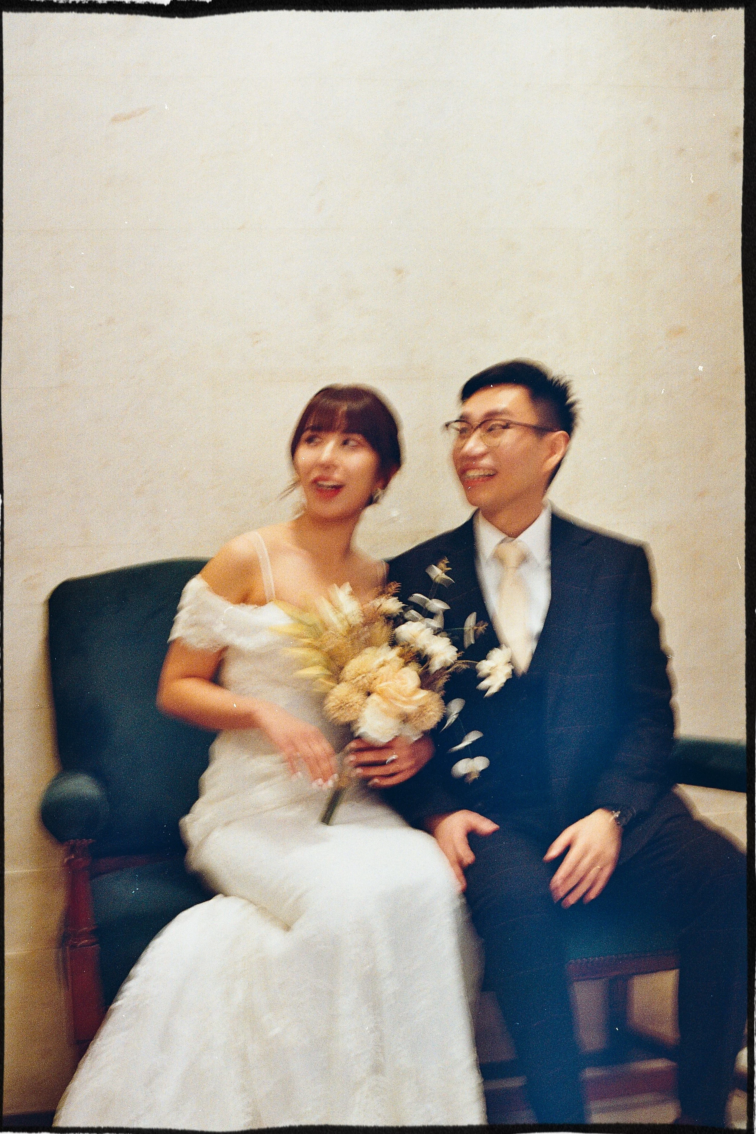 A bride and groom sitting on a bench, smiling and holding a bouquet of flowers, at their wedding reception.