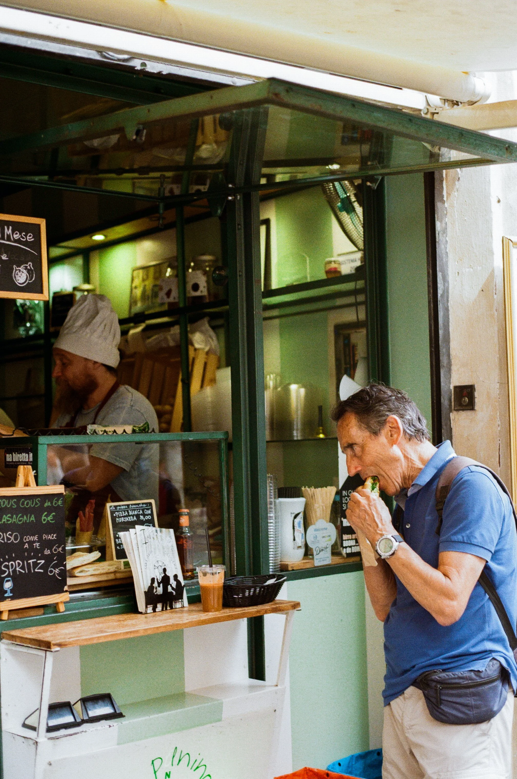 A man in a blue shirt and beige shorts is eating a sandwich outside a small green and white food stand. Inside, a person wearing a chef's hat is preparing food.