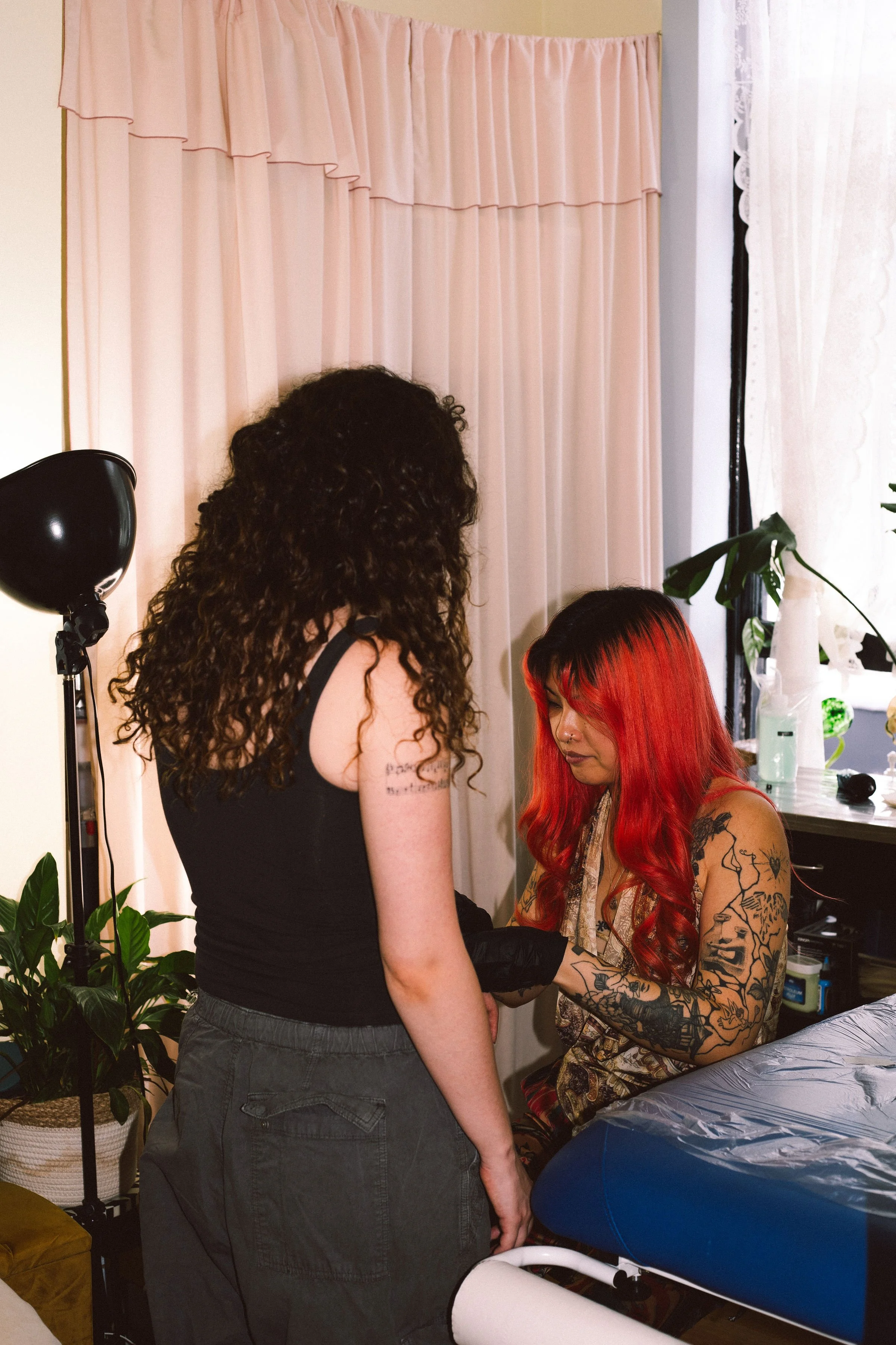 A woman with red hair and tattoos sitting on a chair while another woman with curly dark hair stands nearby. The setting appears to be a tattoo studio with tattoo equipment and various supplies visible.