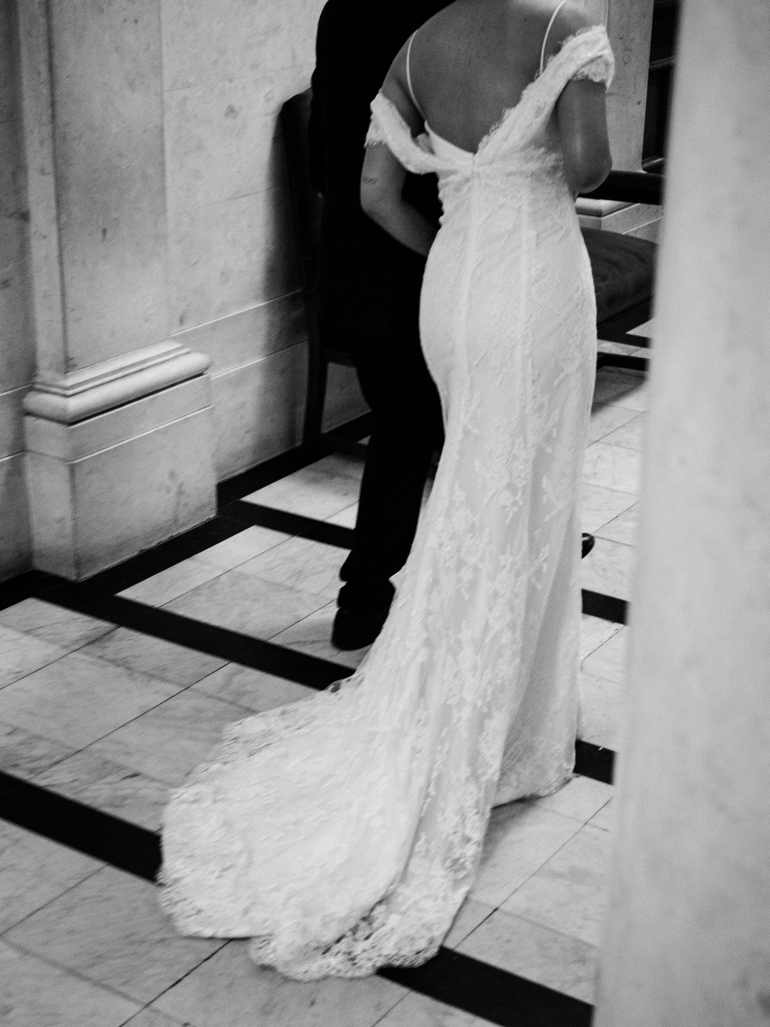 A woman in a wedding dress standing on a tiled floor, facing away, with a man standing behind her. The photo is taken in a mirror, showing their reflections.