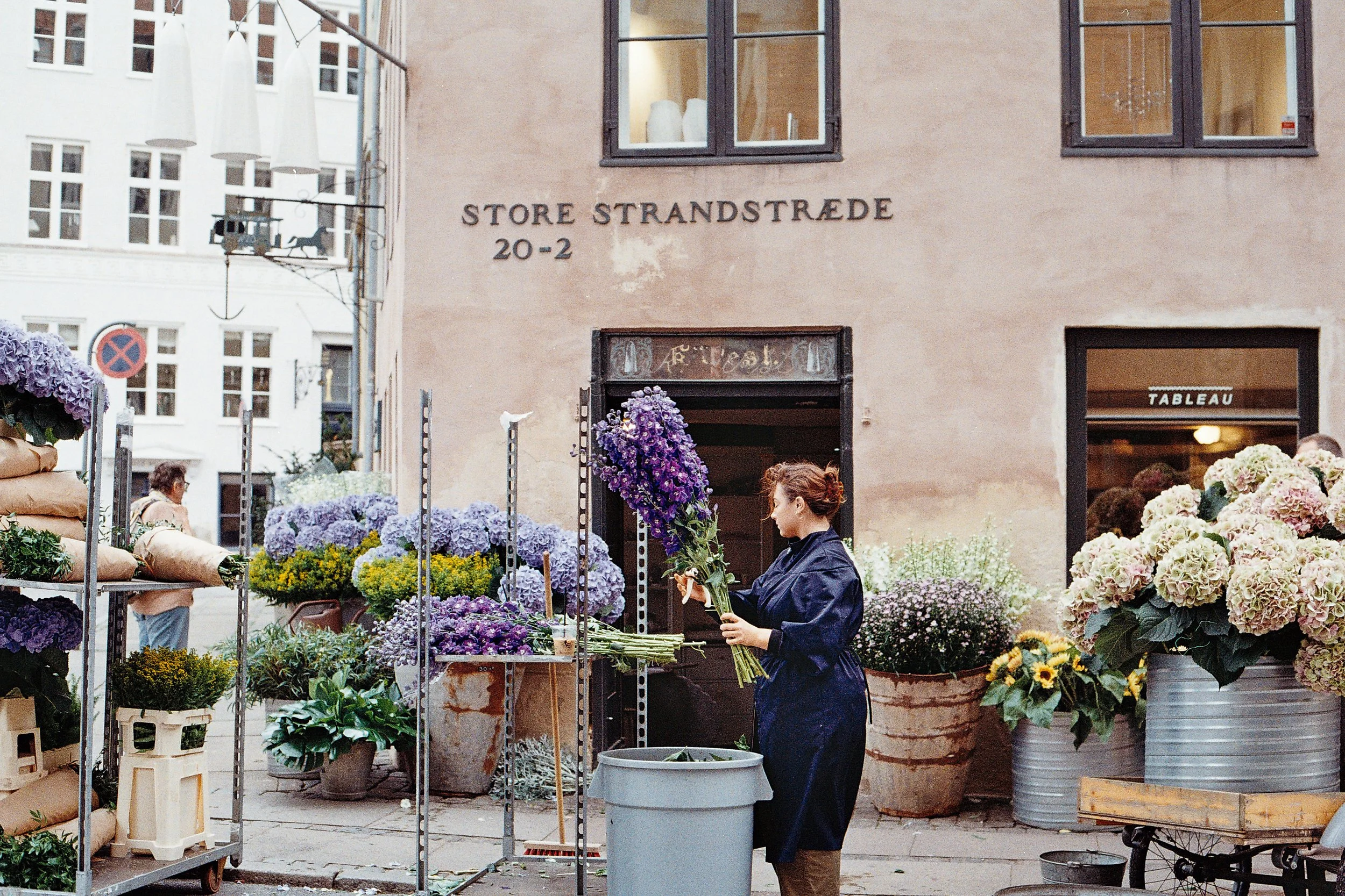 A woman selecting purple flowers at an outdoor flower market in front of a building with sign 'STORE STRANDSTRÆDE 20-2' with large flower arrangements and potted plants around her.