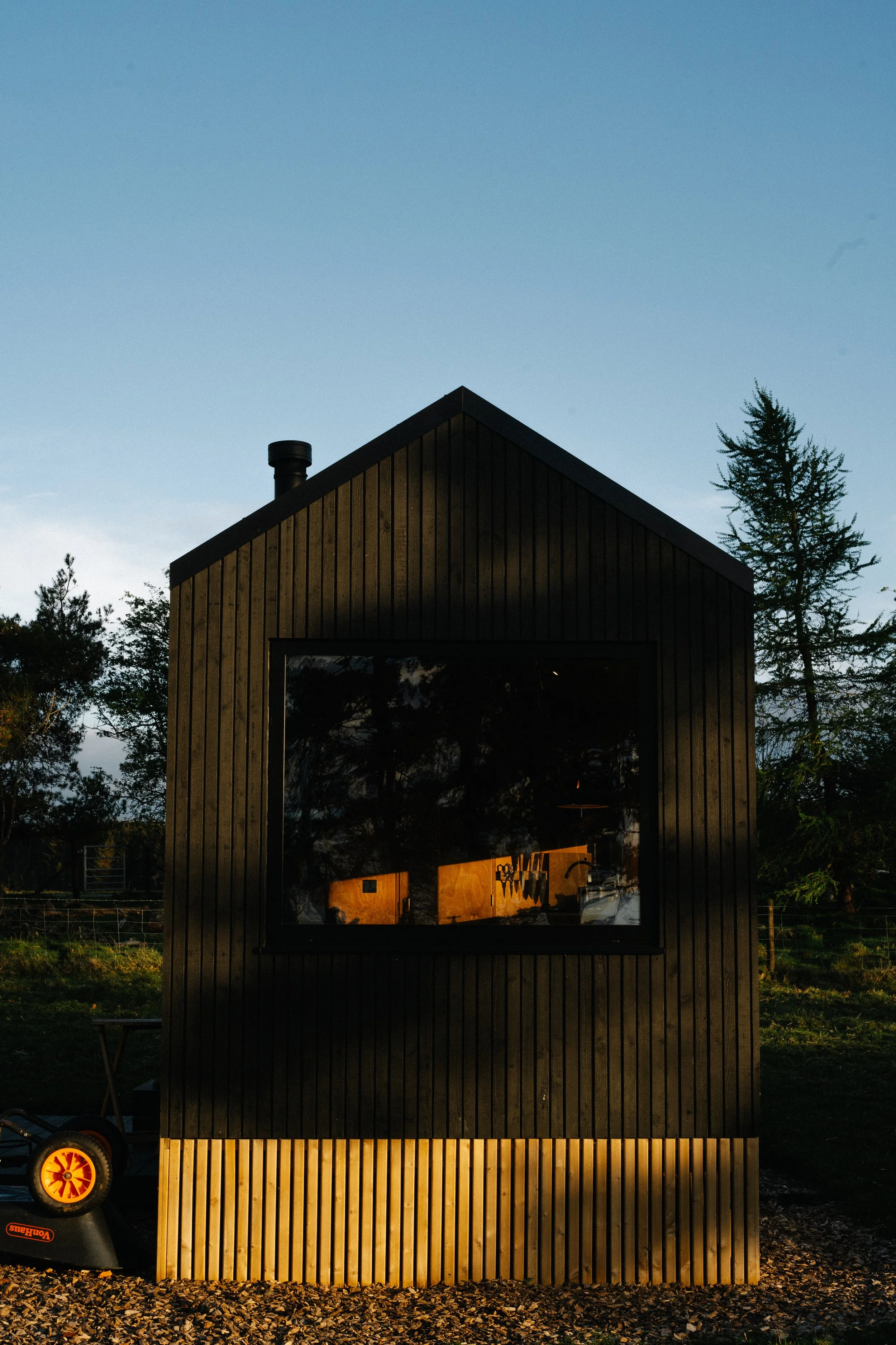 A small black wooden house with a large window reflecting the sunset sky, surrounded by trees and a gravel area at dusk.
