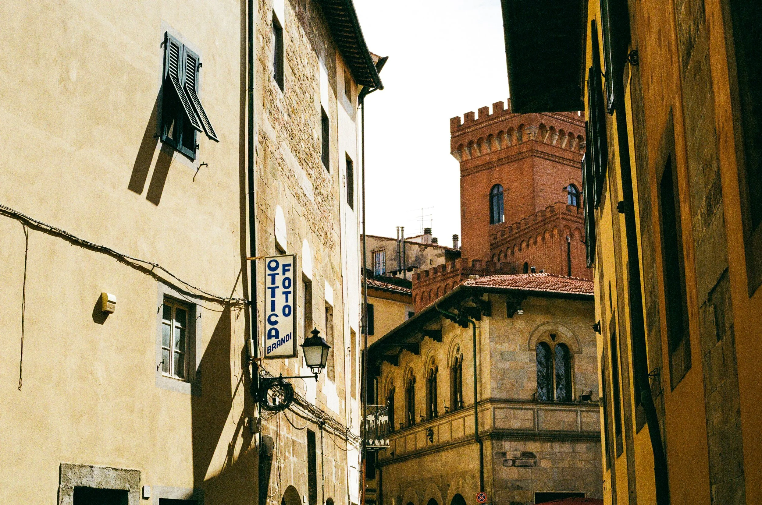 Narrow street lined with old buildings in Italy, featuring yellow, beige, and stone facades, with a prominent red brick tower in the background.