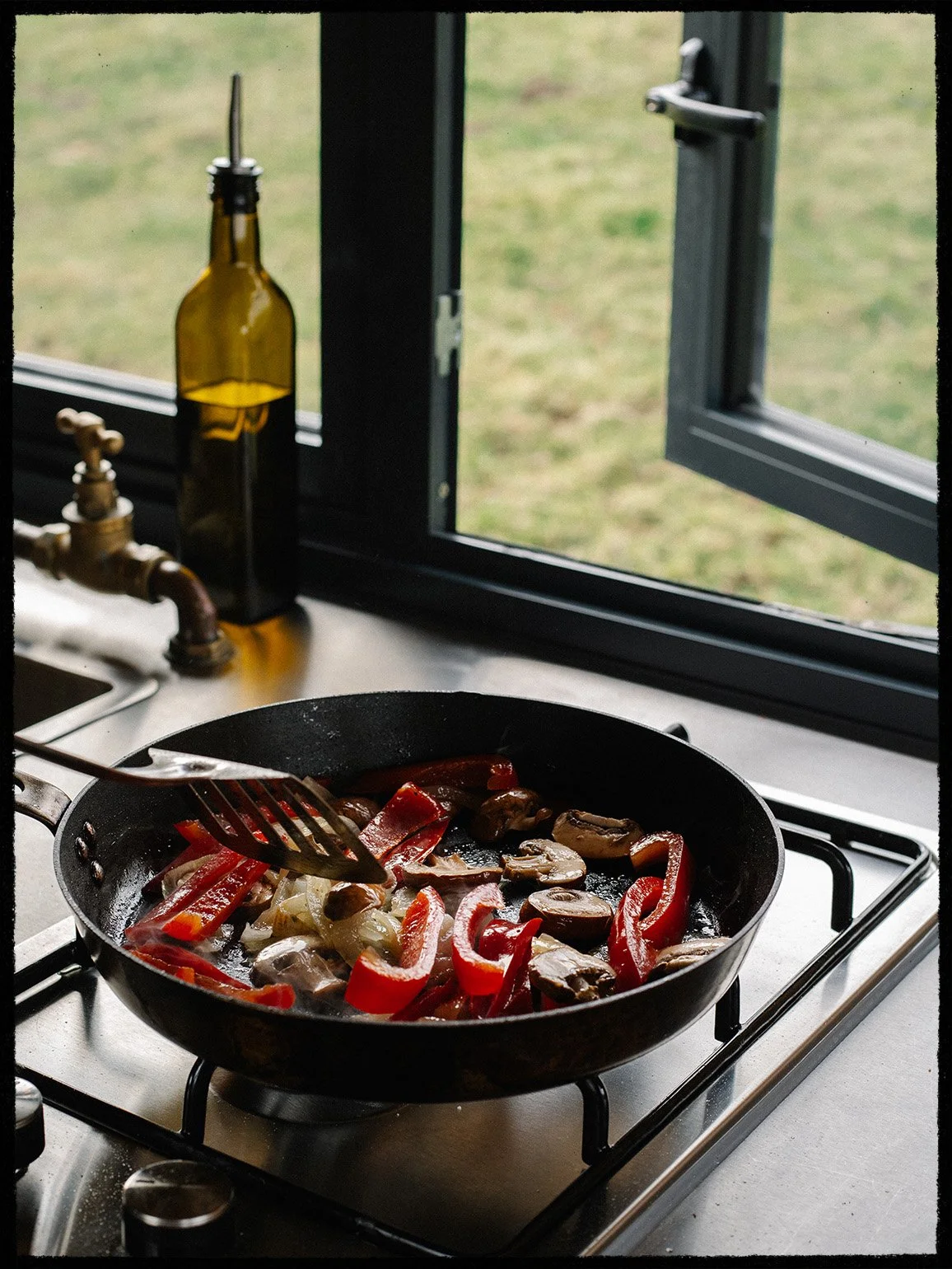 Sautéed red bell peppers, onions, and mushrooms cooking in a black frying pan on a stove with a window in the background.
