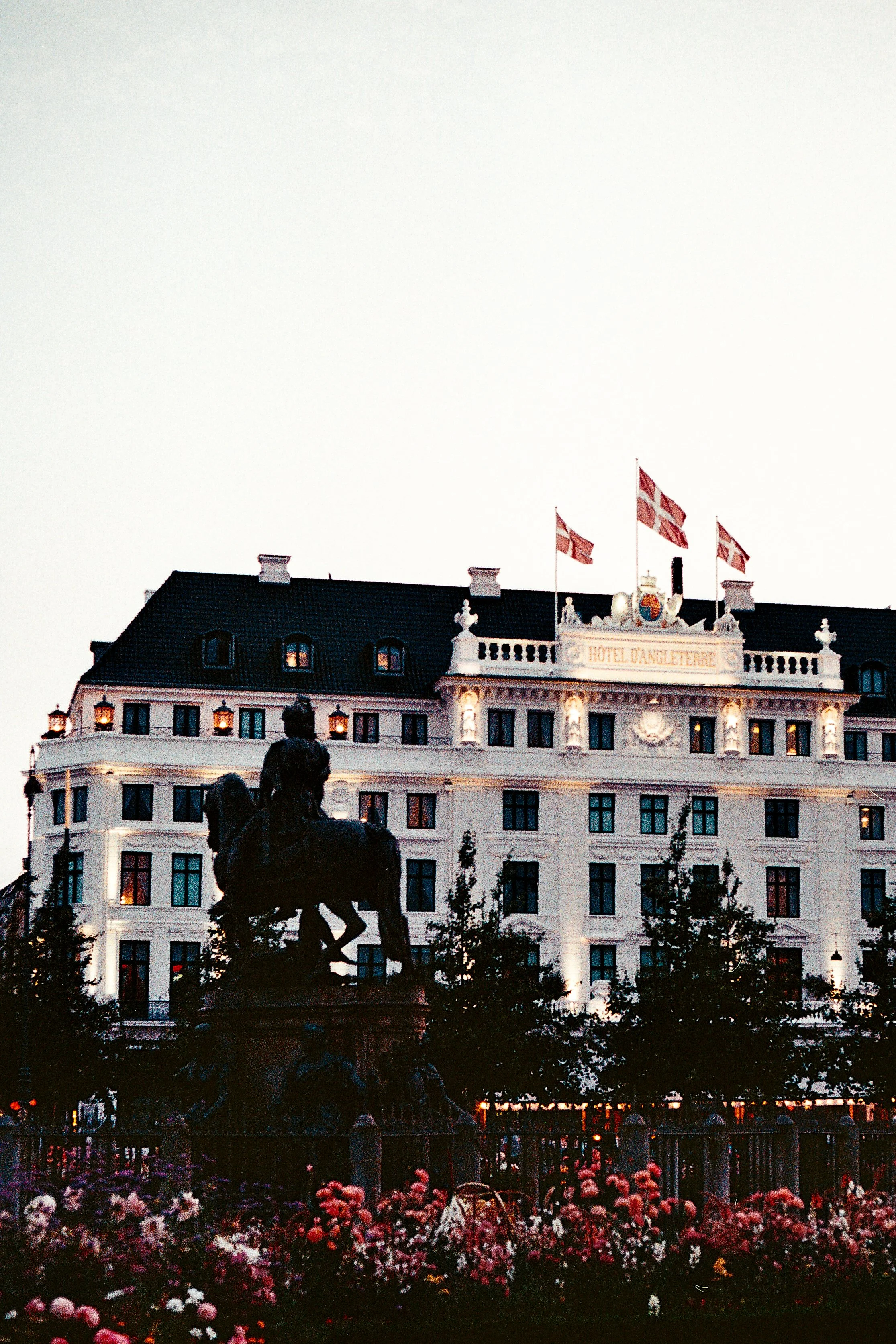 Exterior of Hotel Dangleterre with lit windows and Danish flags, in front of an equestrian statue surrounded by pink and white flowers.