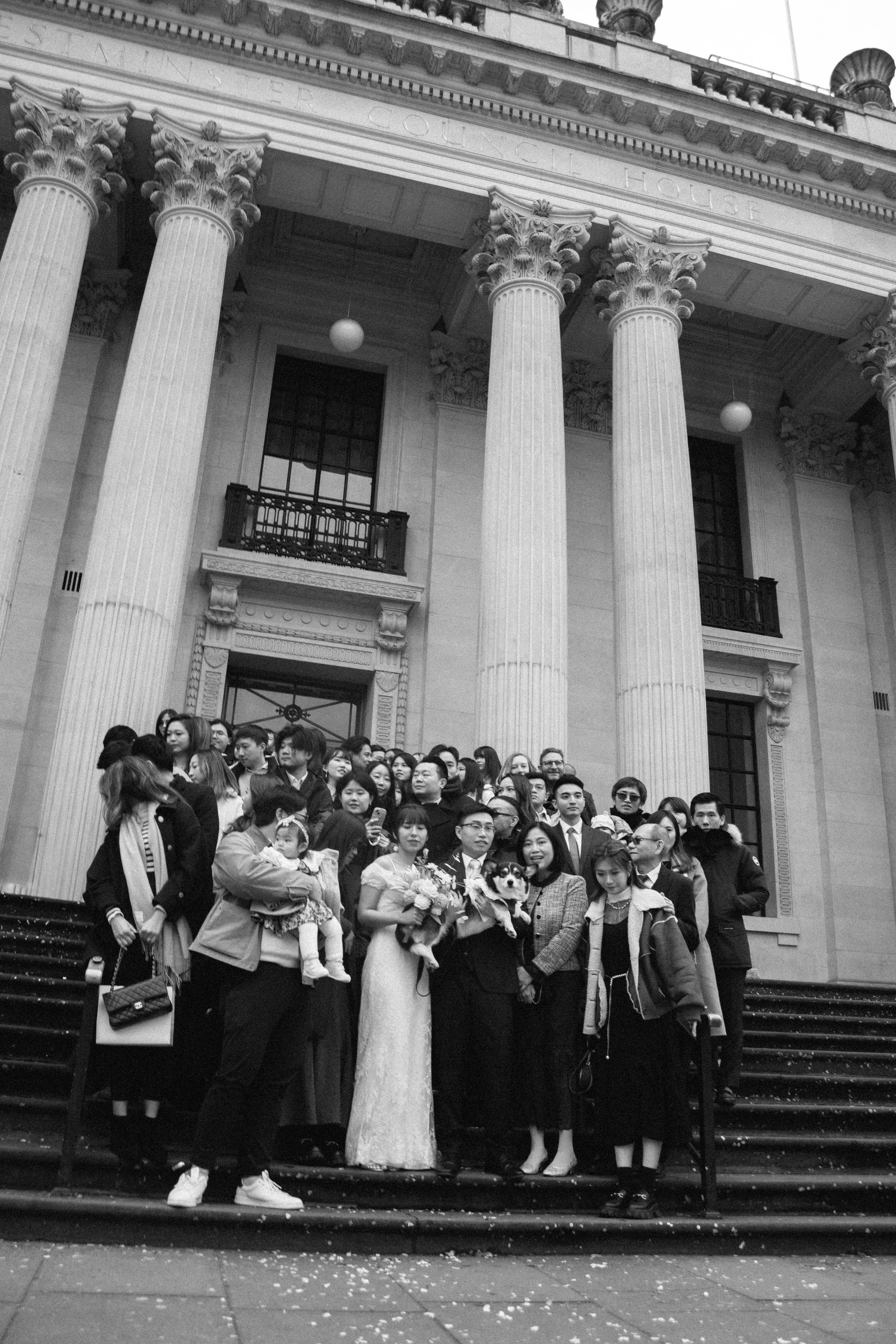 A large group of people, including a bride holding flowers and a groom with a dog, standing on the steps of a historic building with tall columns and ornate architecture.
