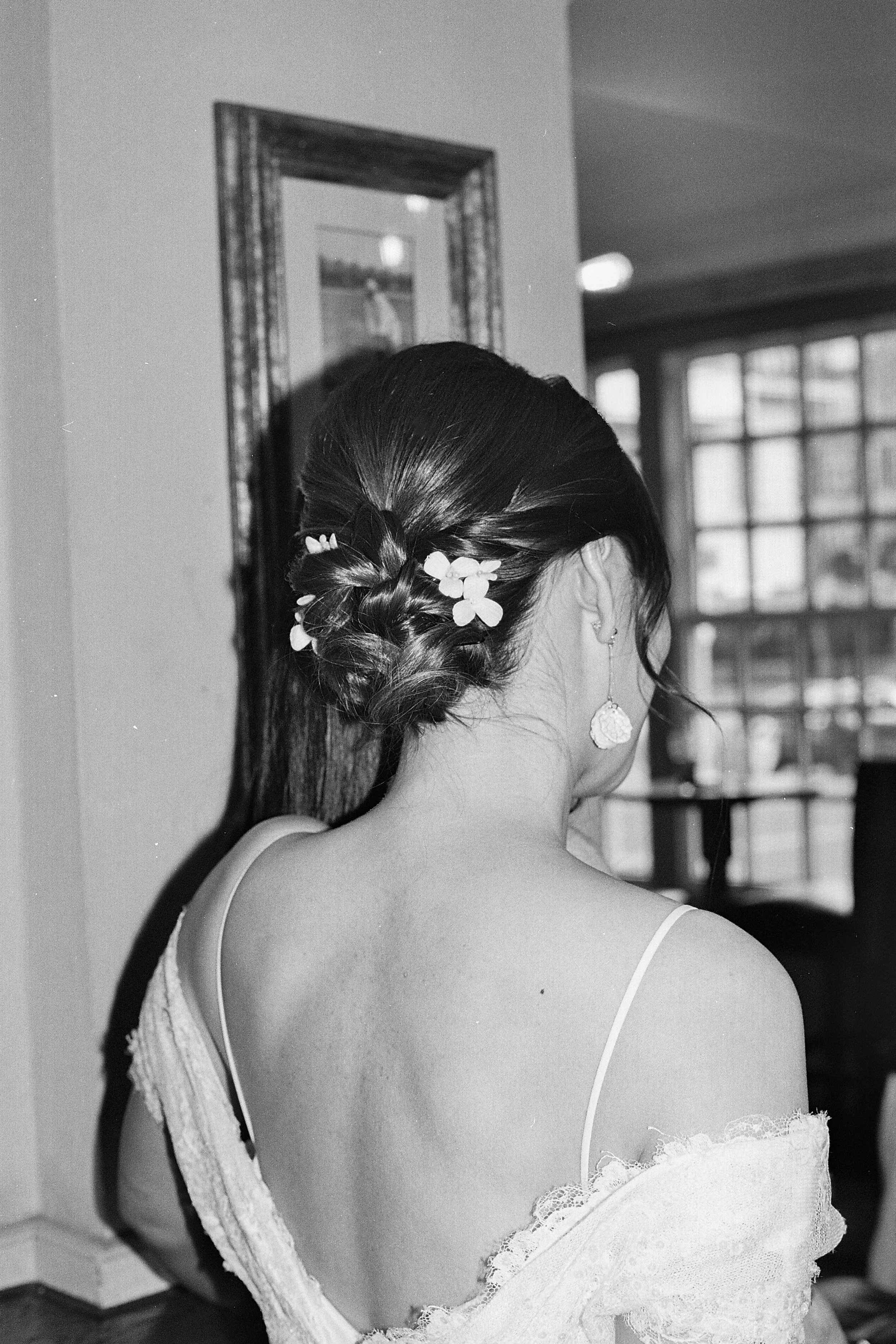 Back of a woman's head with an elegant braided hairstyle decorated with small flowers, wearing a sleeveless dress with lace straps, earrings, and a tattoo on her shoulder, inside a room with large windows.