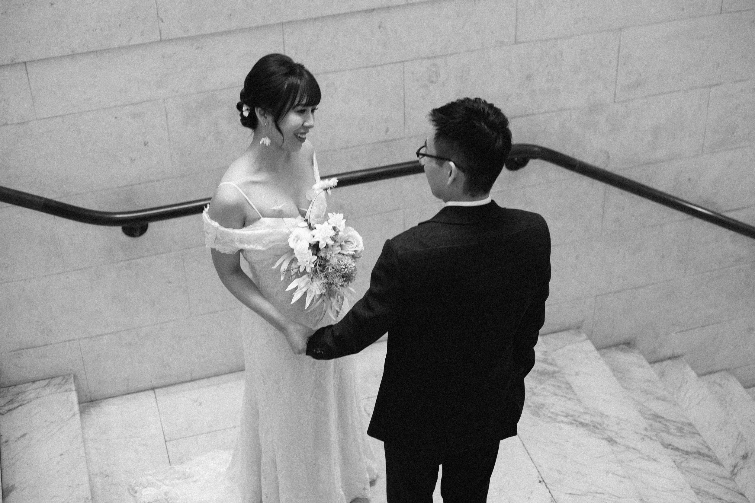A bride and groom holding hands on a staircase, smiling at each other, with the bride holding a bouquet of flowers.