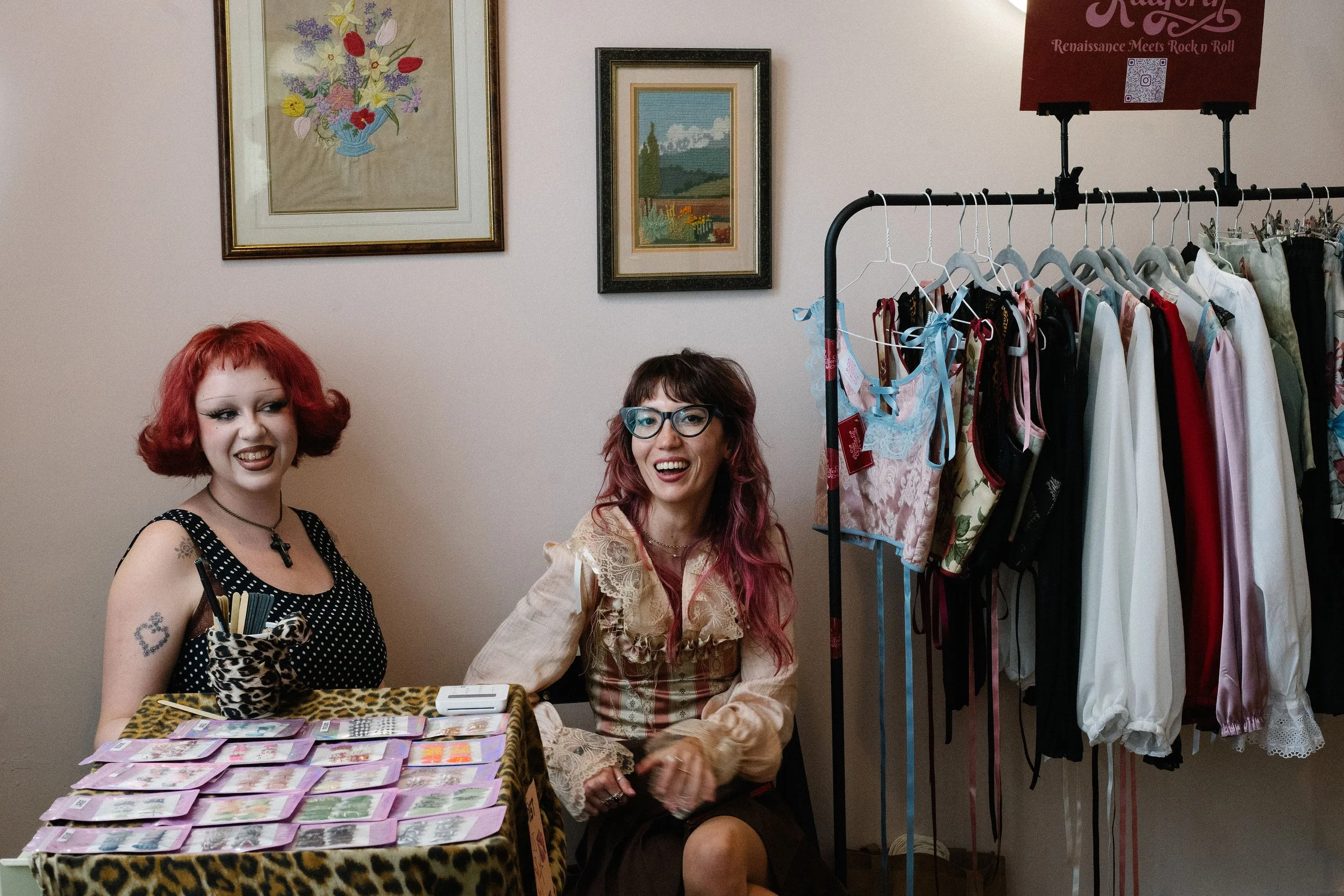 Two women sitting at a table filled with pink tickets or cards, with a rack of lingerie and clothing behind them. The woman on the left has red hair, is wearing a black polka dot dress, and has tattoos, while the woman on the right has pink and brown hair, glasses, and is wearing a beige blouse with lace details, smiling and laughing.