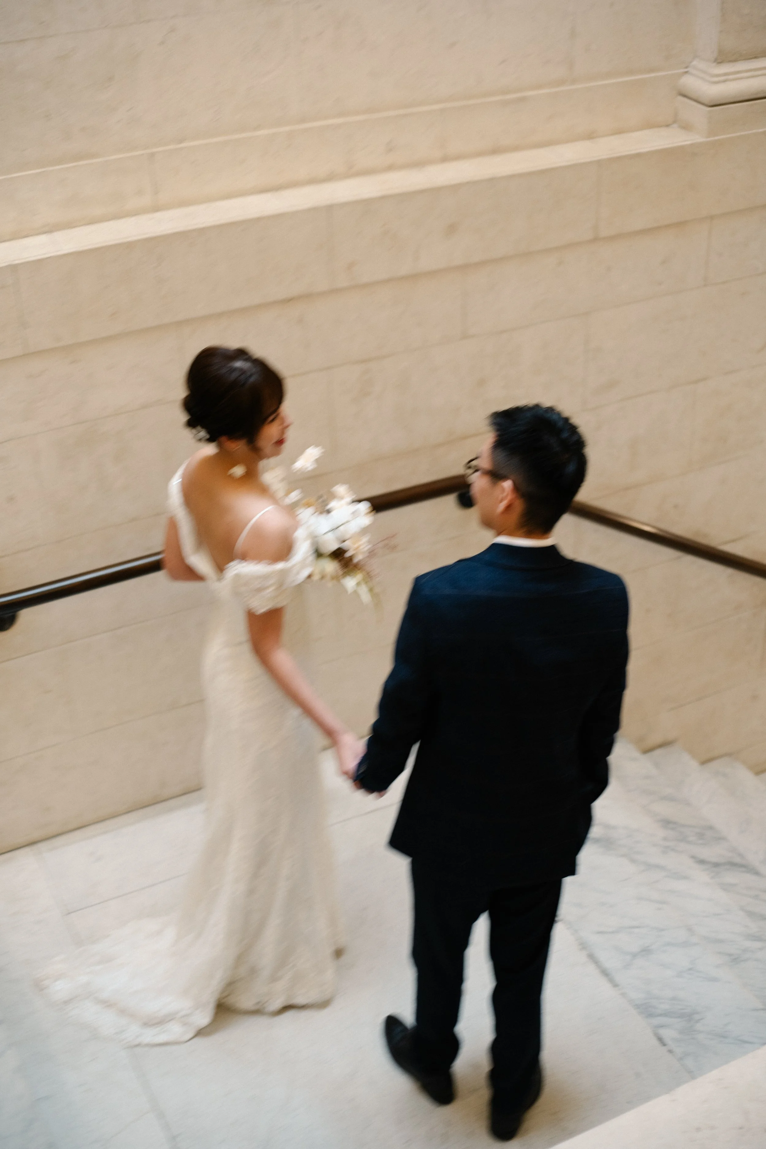 A bride and groom holding hands on a staircase in an elegant indoor setting, with the bride holding a bouquet of white flowers, wearing a white wedding dress and the groom in a dark suit.