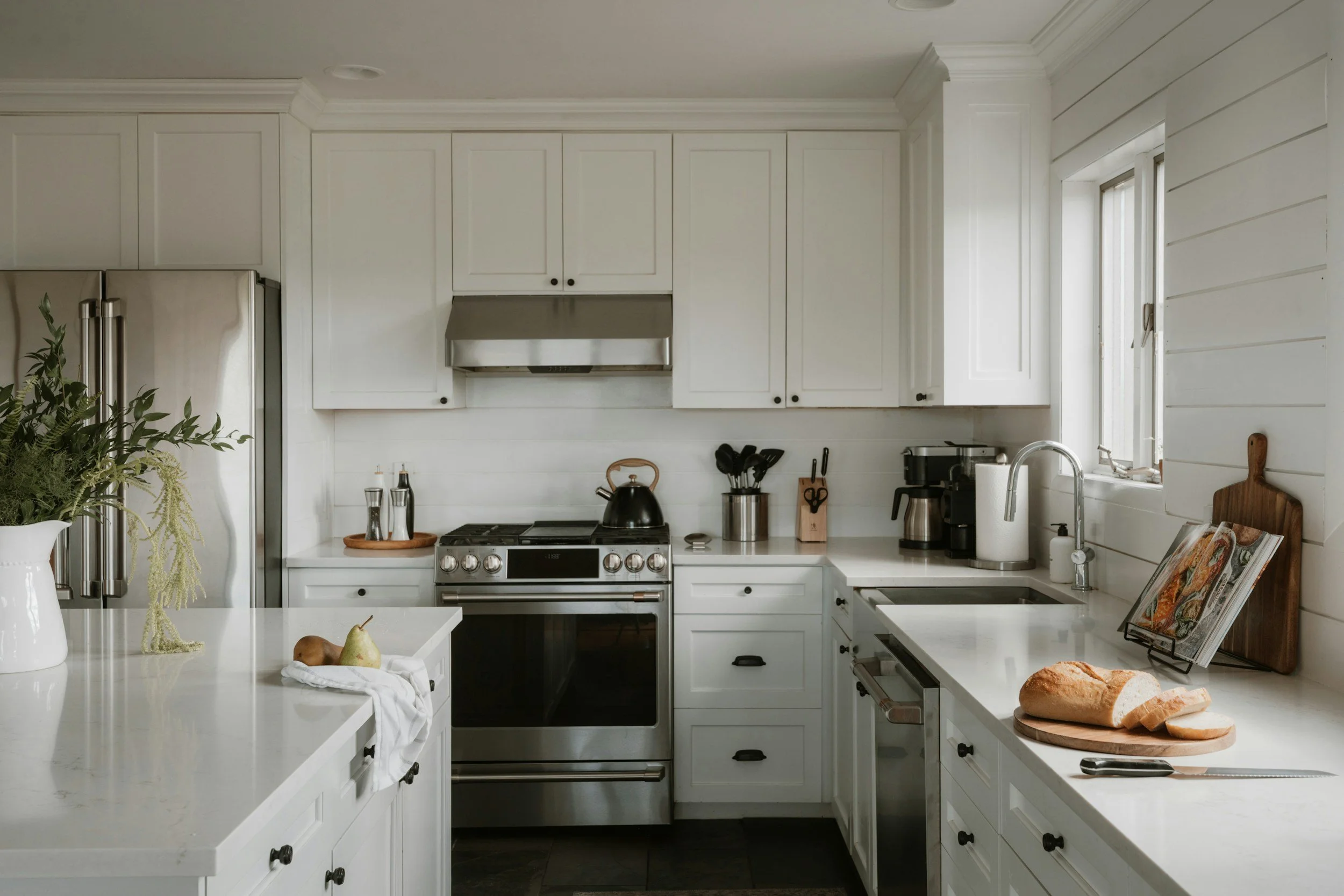 White kitchen with modern appliances, including stainless steel refrigerator, oven, stove, and coffee machine, with white cabinets and countertops, a window, and a loaf of bread on a wooden cutting board.