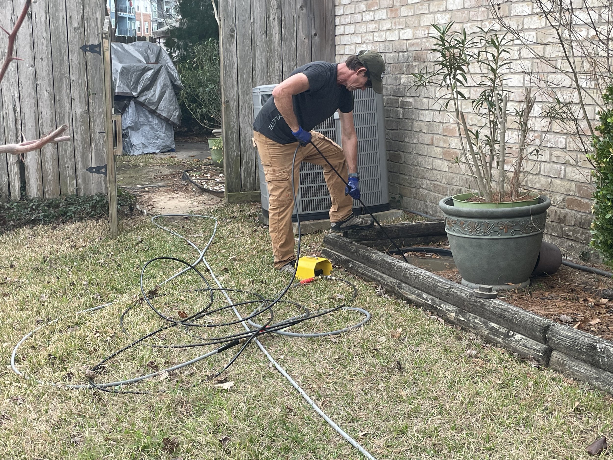 Man working outdoors with tools, standing next to brick wall and plants, with electrical cords on grass.