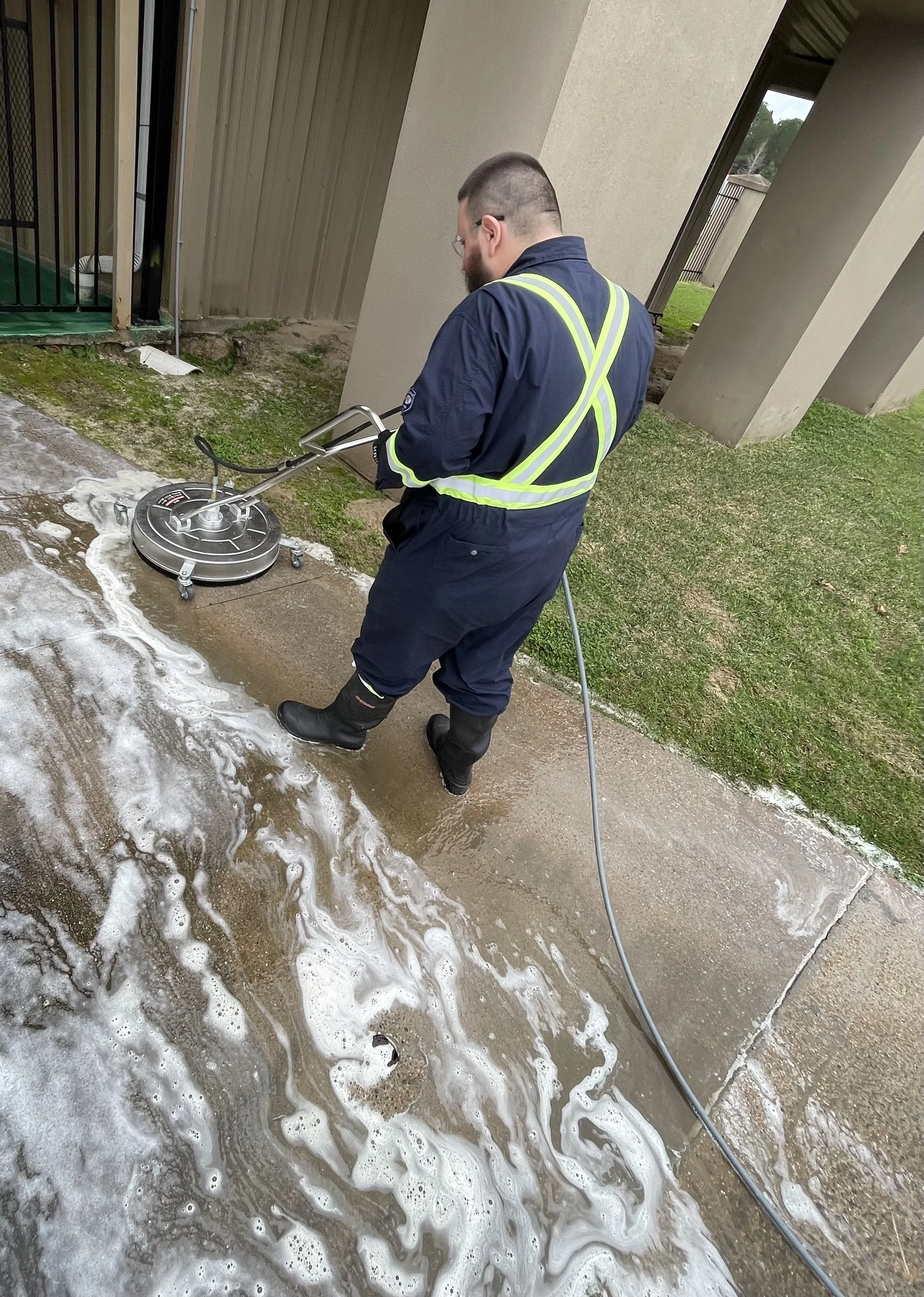 A man wearing a navy uniform with reflective yellow-green straps uses a pressure washer to clean a concrete sidewalk near a building, with soap suds and water on the ground.