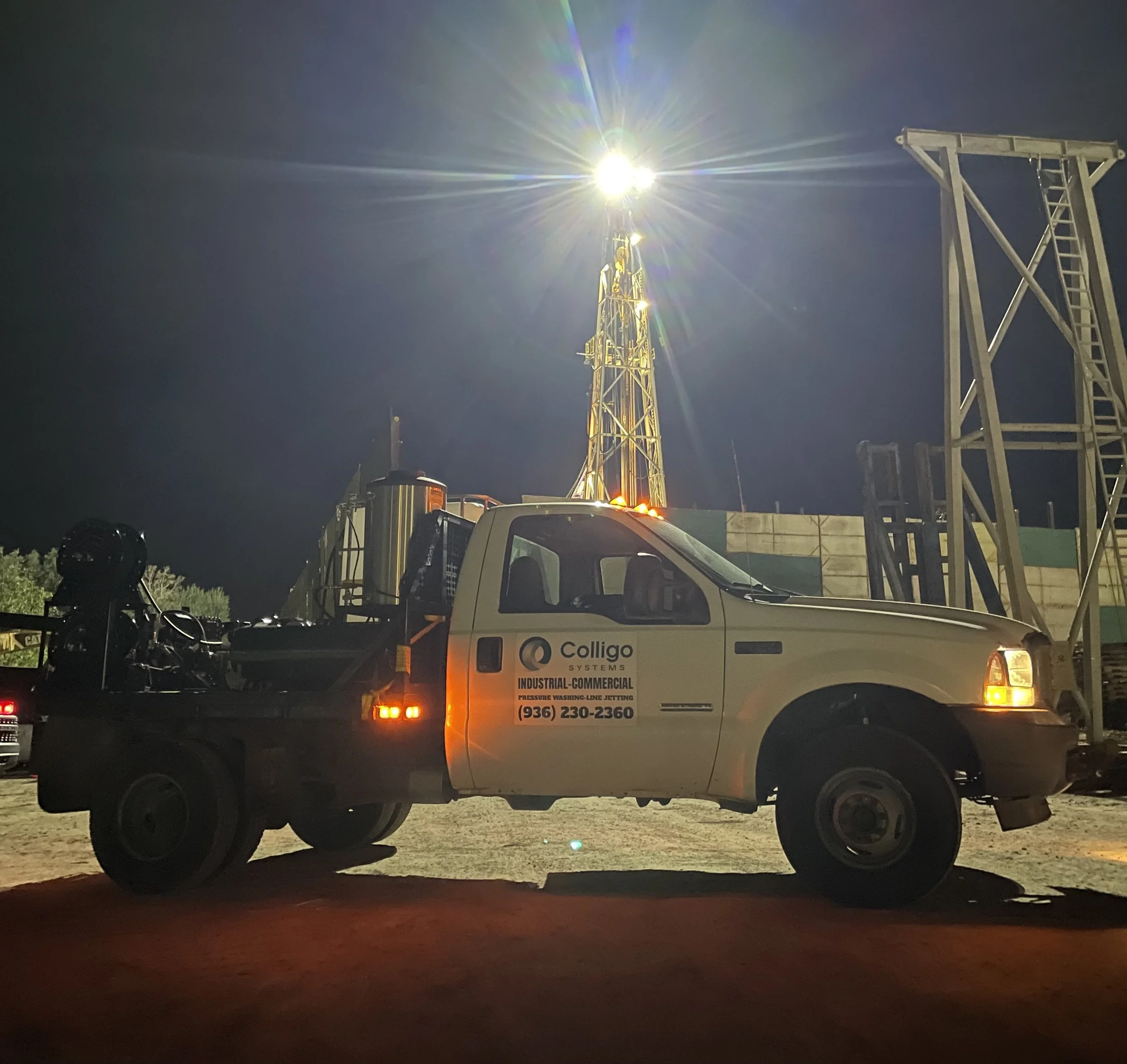 A nighttime image of a service truck with a company logo for Colligo Systems, parked at a construction or industrial site with a tall lighting tower illuminating the area. The scene shows equipment and structures related to industrial or construction work.