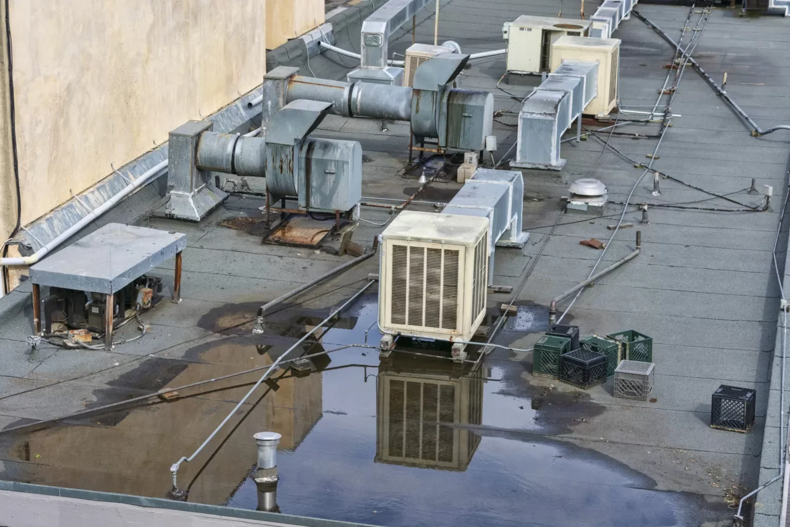 Rooftop with multiple HVAC units, pipes, electrical wires, and a small puddle of water reflecting some equipment.
