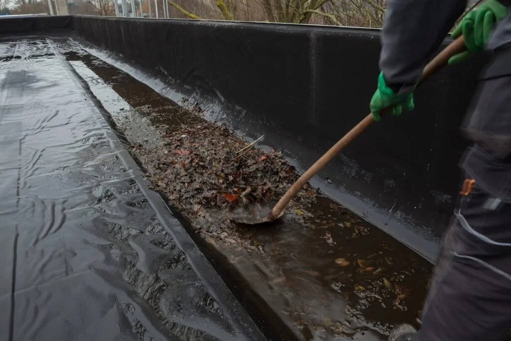 Person wearing green gloves shoveling leaves and debris from a black rain gutter or drainage system.
