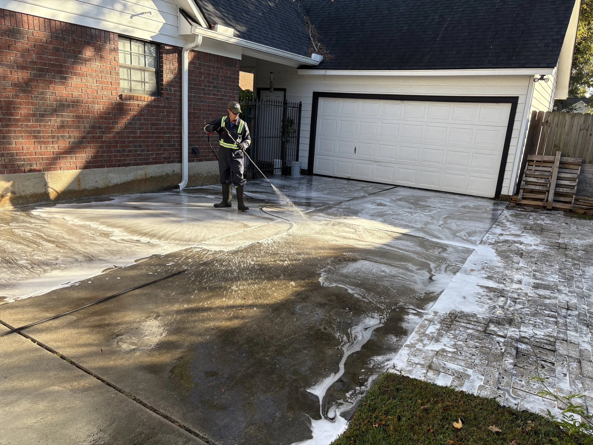 A person wearing protective gear power washing a concrete driveway outside a brick house with a white garage door.