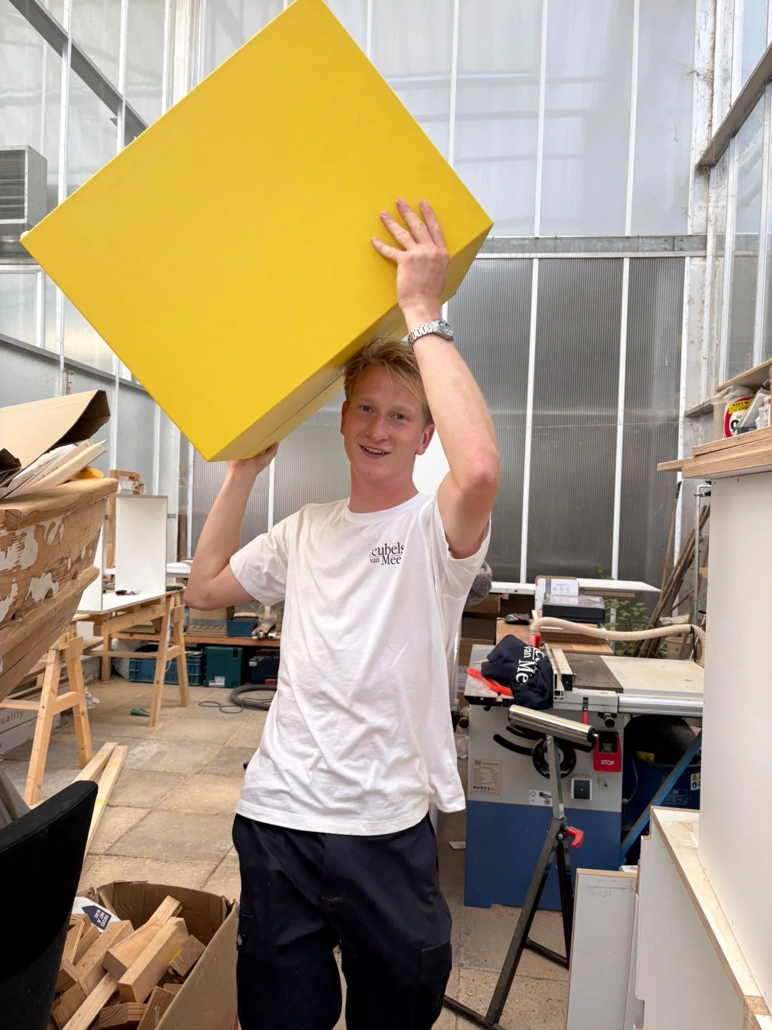 Een jonge man in een werkplaats tilt een grote gele kubus boven zijn hoofd.