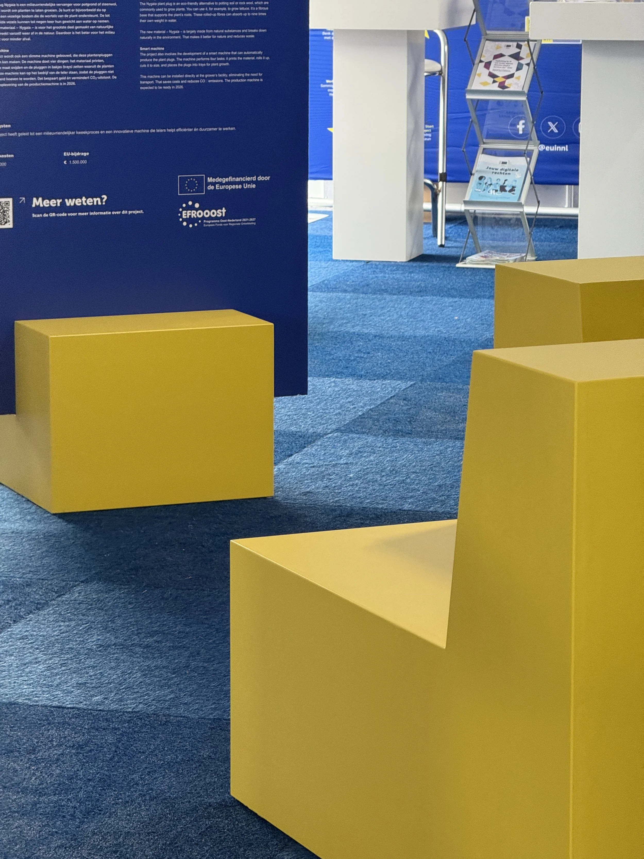 Een overzicht van een informatieset-up met gele stoelen en blauw paneel met teksten en logos, inclusief informatie over het project en de Europese Unie.