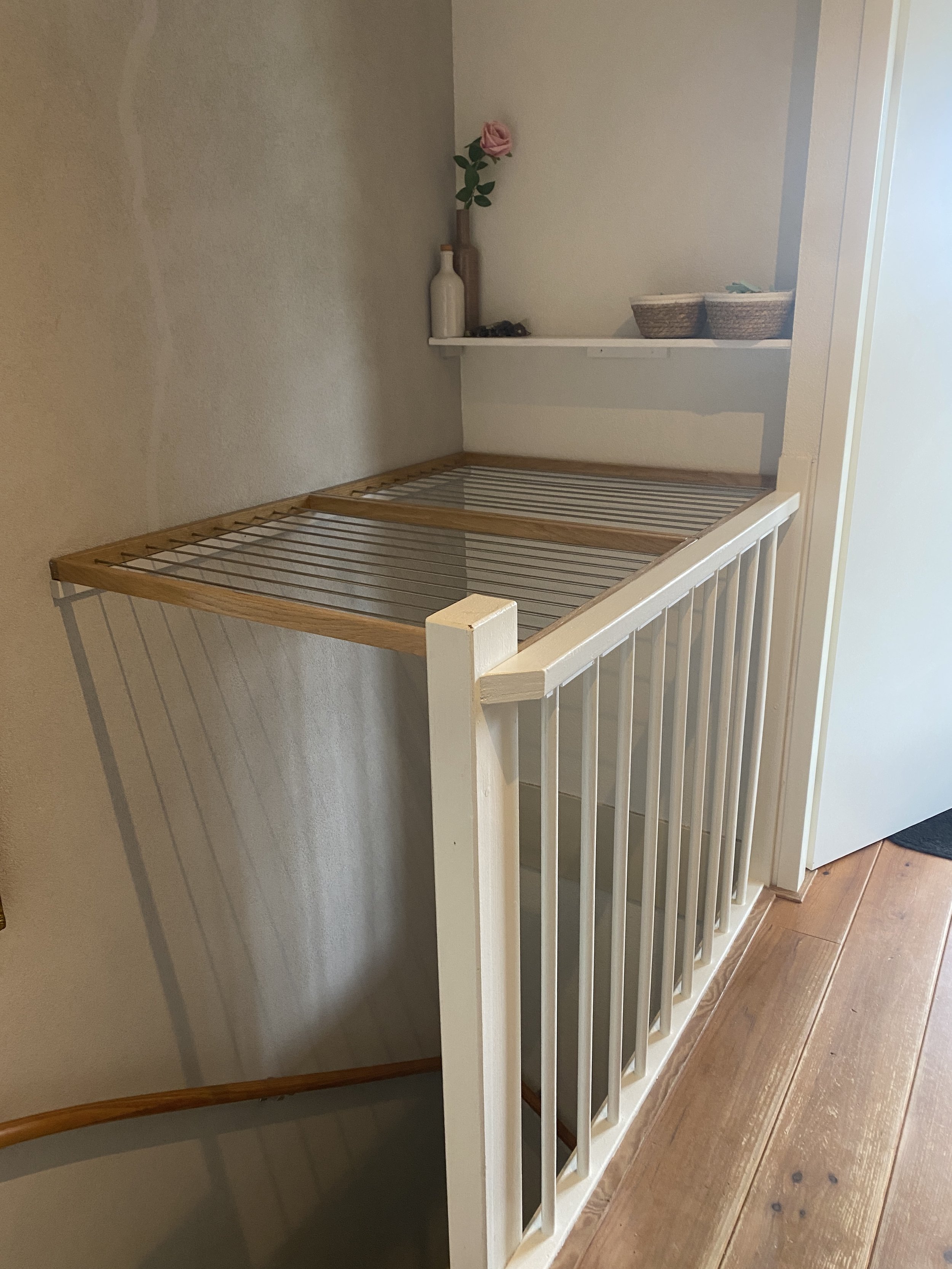 Bovenaan een wit en houten wasrek met een glazen blad, naast een kleine witte muur met een smalle plank met decoratieve vazen en een bloem, gelegen bij een houten vloer.