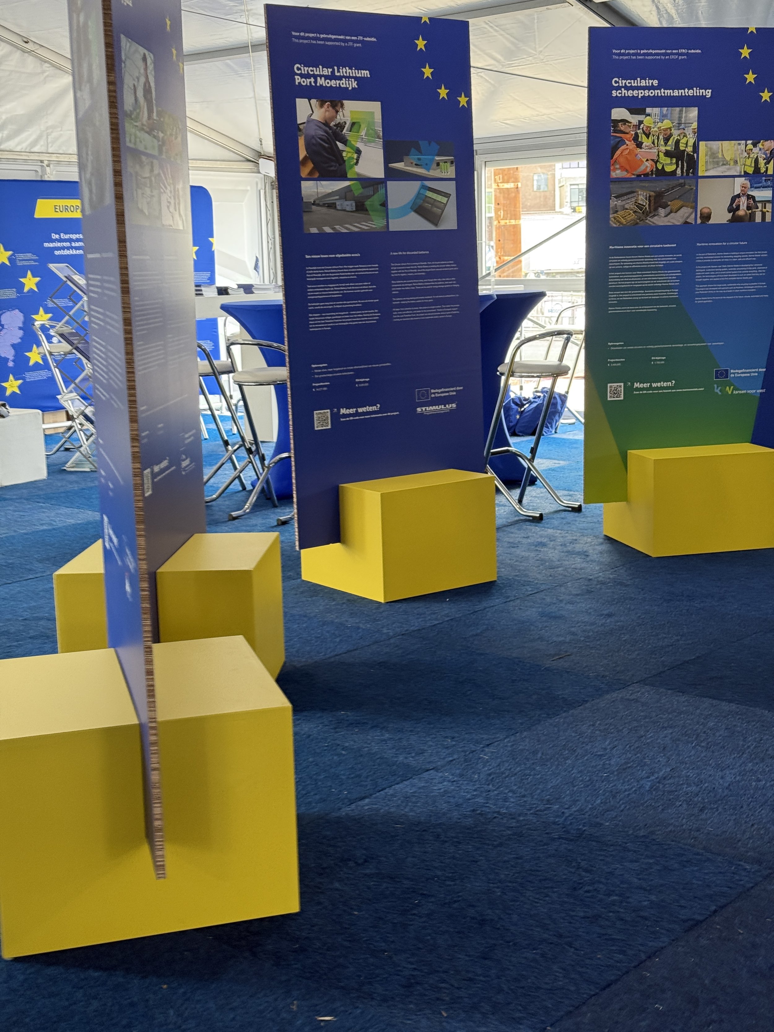Informatieborden met teksten en foto's over circulariteit en milieuvriendelijke projecten, geplaatst op gele blokken in een tent met blauwe vloer en blauwe achtergrond.