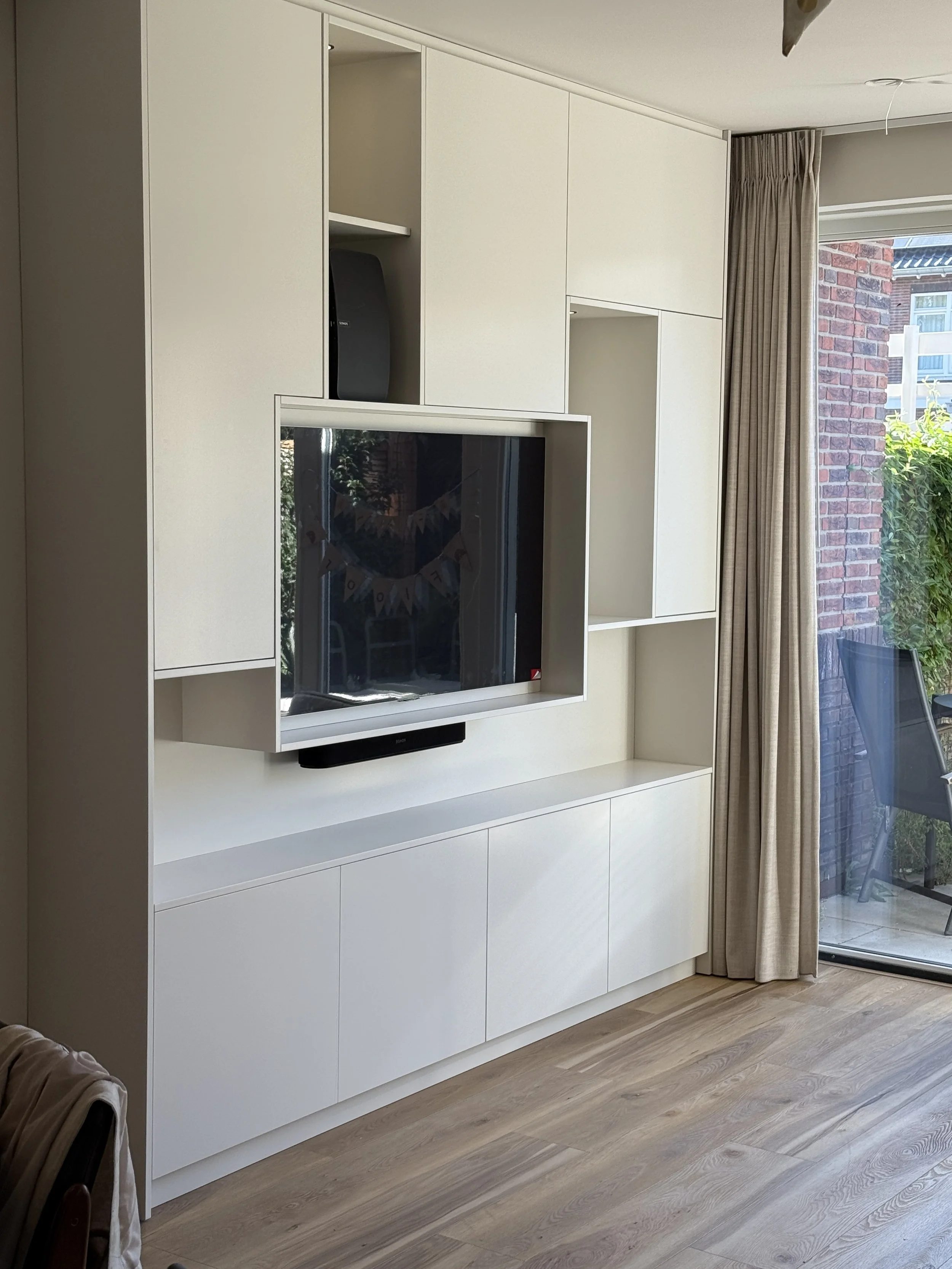 Witte muur met ingebouwde kast en tv, grote raampartij met beige gordijn, terras met tuinstoel buiten.