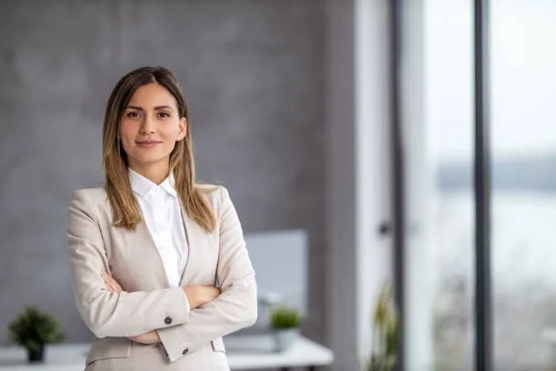 Confident woman in business attire standing with arms crossed in modern office with large windows.
