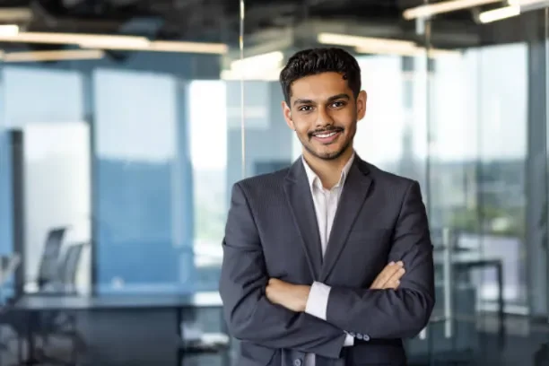 A smiling man in a dark suit with arms crossed standing in a modern office with glass walls.