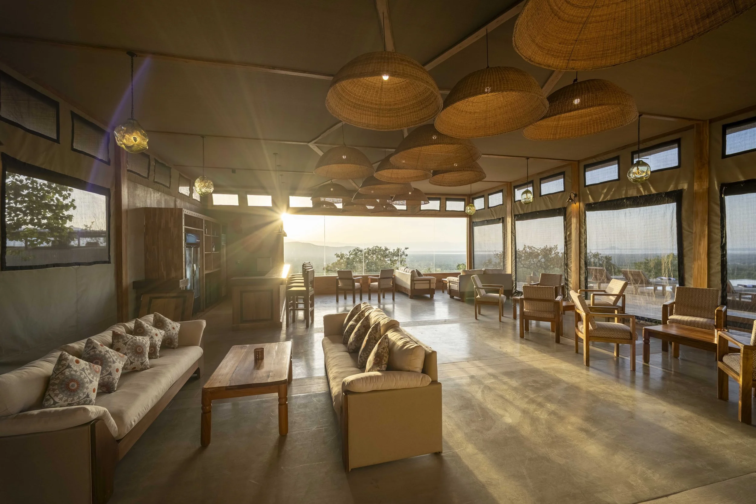 A spacious sunlit lounge area with large windows, wicker ceiling lights, and various seating options including sofas and chairs, overlooking a scenic view of trees and water.