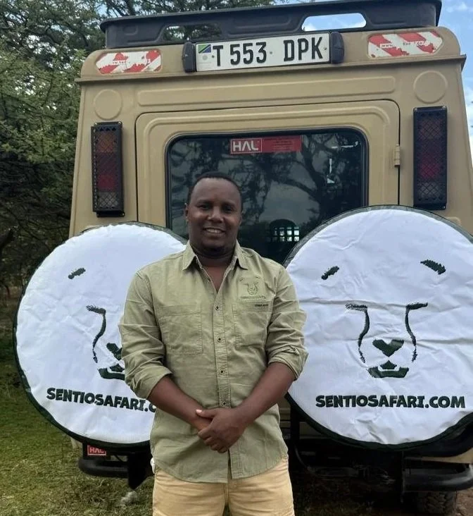 A man standing in front of a safari vehicle with sentiosafari.com logos on the tires, smiling and wearing a beige shirt, outdoors with trees in the background.